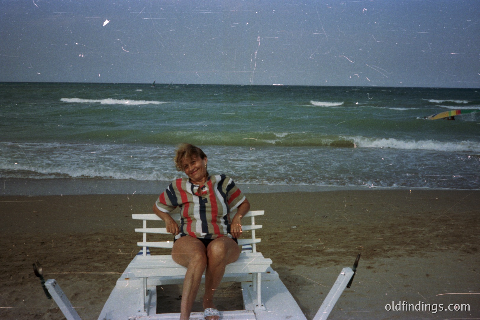 A woman sits on a white, folding beach chair facing the camera. She wears a patterned, short-sleeved top and shorts. Behind her, a beach stretches to the turquoise sea. The image exhibits visible film grain and light leaks. Likely captured in the 1970s or 1980s.