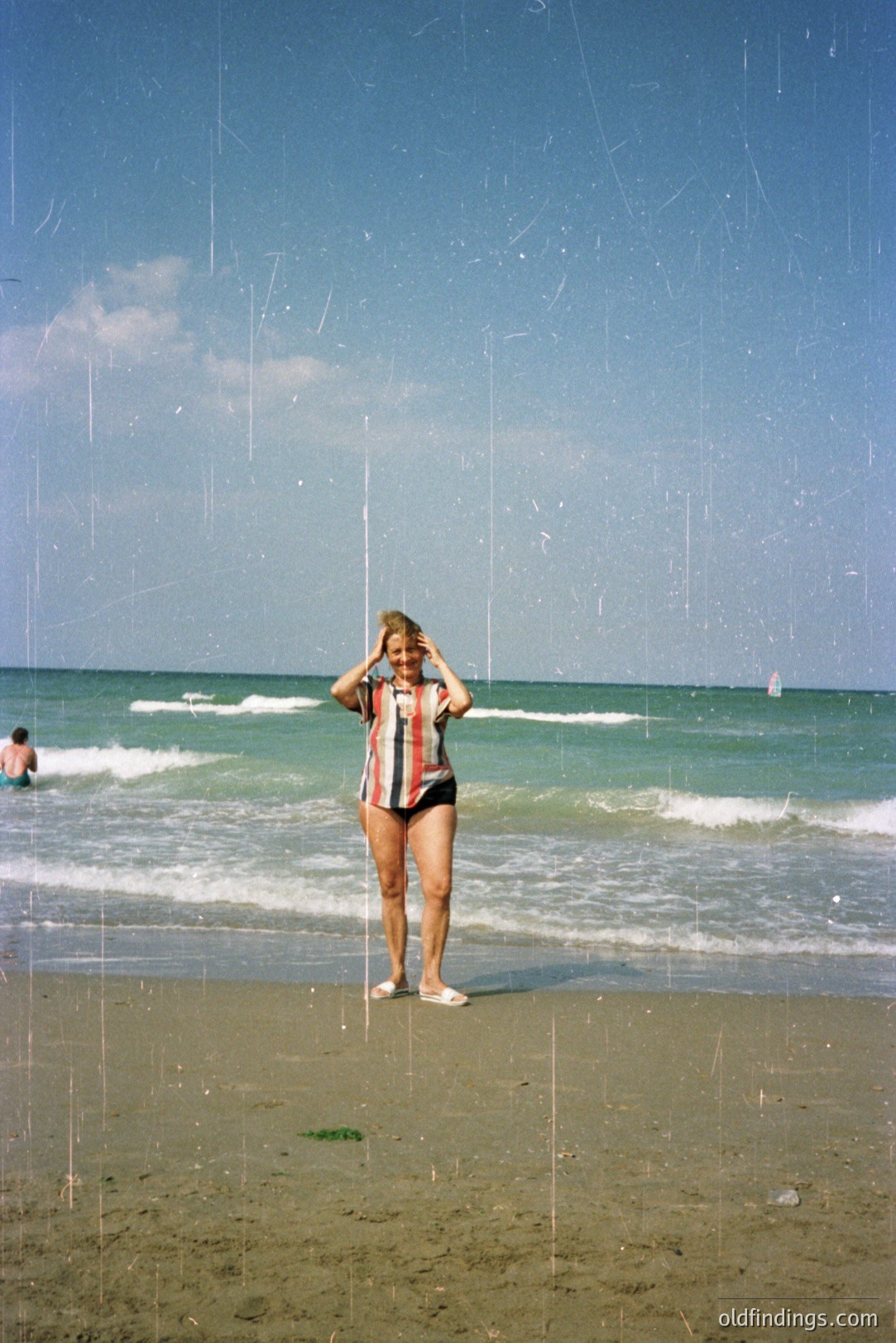 A young woman in a vintage, patterned swimsuit and short sleeves stands on a sandy beach, appearing to pose for the camera. Gentle waves meet the shore, with a lone swimmer visible in the distance. Strong vertical scratches mark the image, suggestive of aged film. Likely 1970s coastal leisure.
