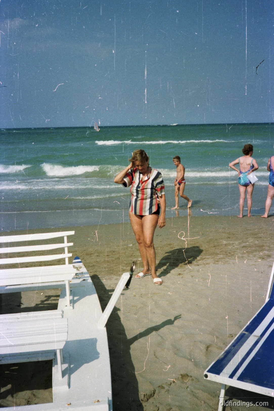 A woman in a patterned swimsuit stands on a sandy beach, casting a shadow near white lounge chairs. Visible in background are beachgoers and a bright, expansive ocean. Likely a vacation snapshot from the 1970s or early 80s.