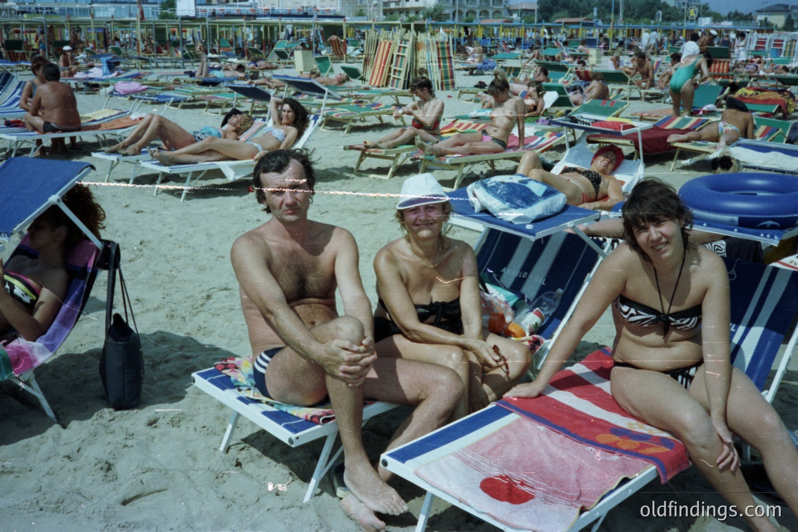 Three people seated on beach chairs at a crowded sandy shoreline. Visible details include patterned towels, dark swimsuits, and a striped beach umbrella. Likely a tourist destination, possibly in Eastern Europe, 1970s. Stock potential for depicting leisure and travel.