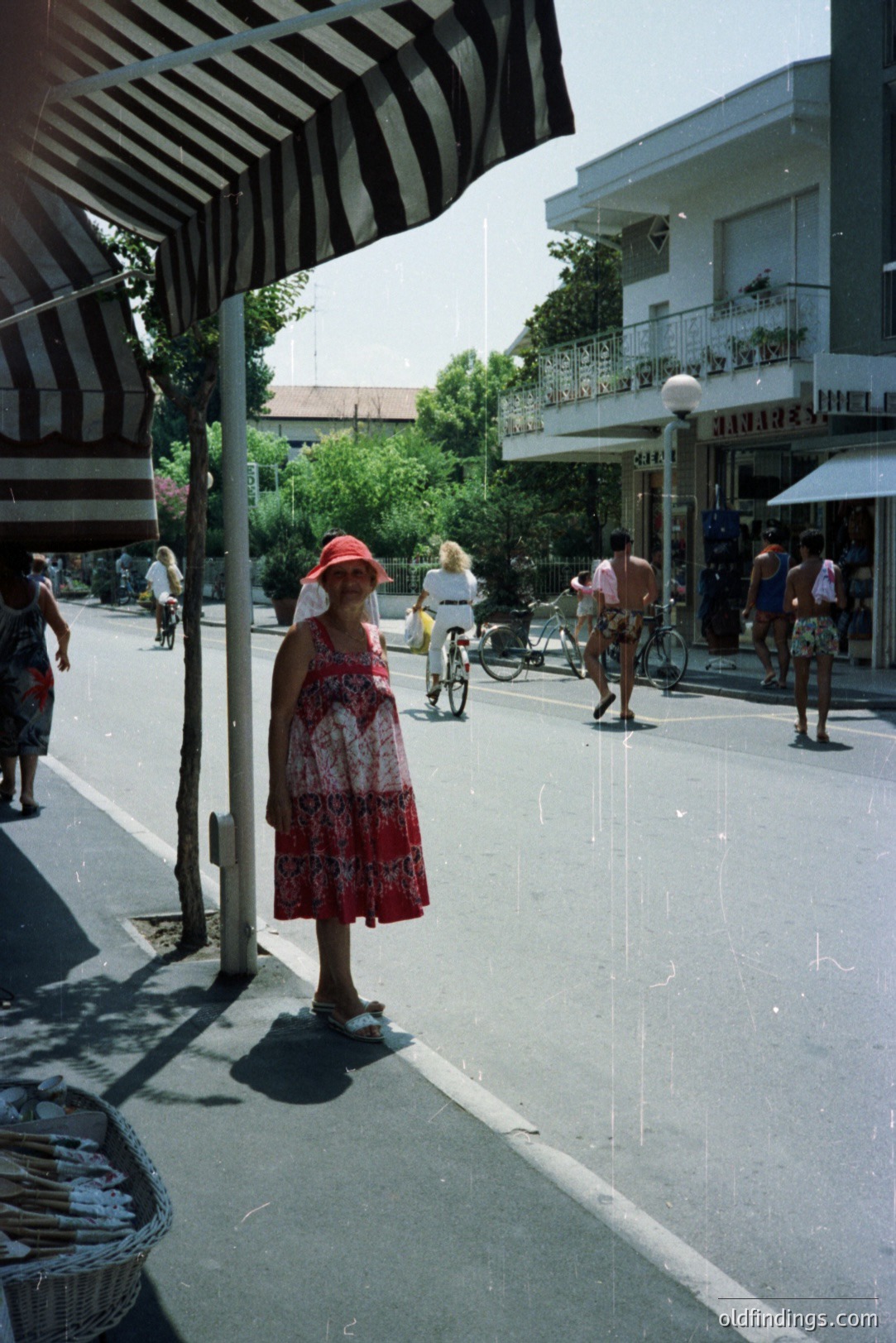 A woman in a floral dress & straw hat stands near a striped awning on a sun-drenched street lined with shops and bicycles. Men in swimwear stroll by. Suggests a seaside resort, possibly in Bulgaria or a similar Eastern European coastal location. Appears to be from the 1980s, judging by the clothing styles and film quality.