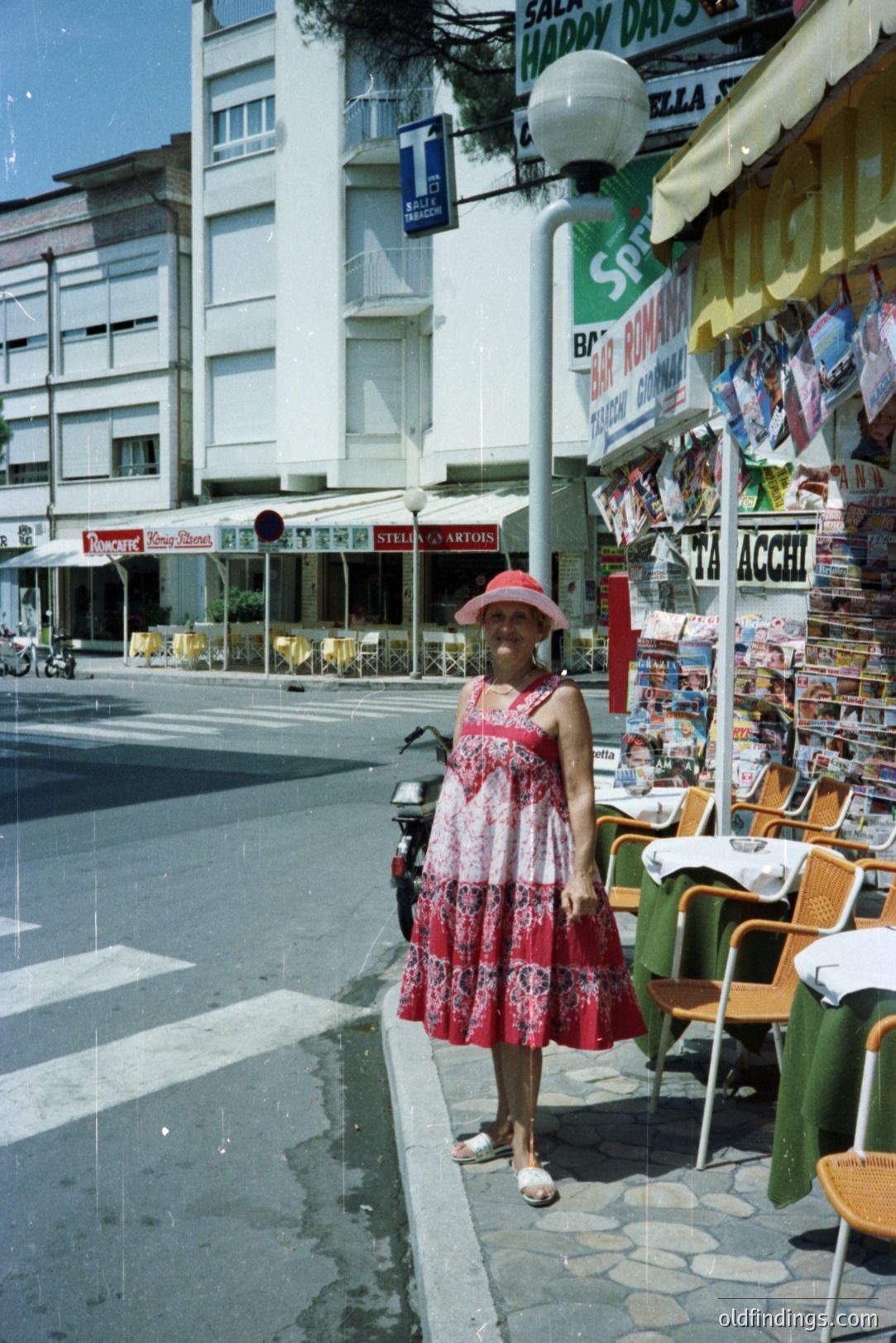 A woman in a floral dress and straw hat stands near a sidewalk cafe, a bustling street scene visible behind her. The photo exhibits a vintage aesthetic, likely from the 1970s. Likely a seaside resort town with commercial storefronts. Appears to be a travel souvenir stand.