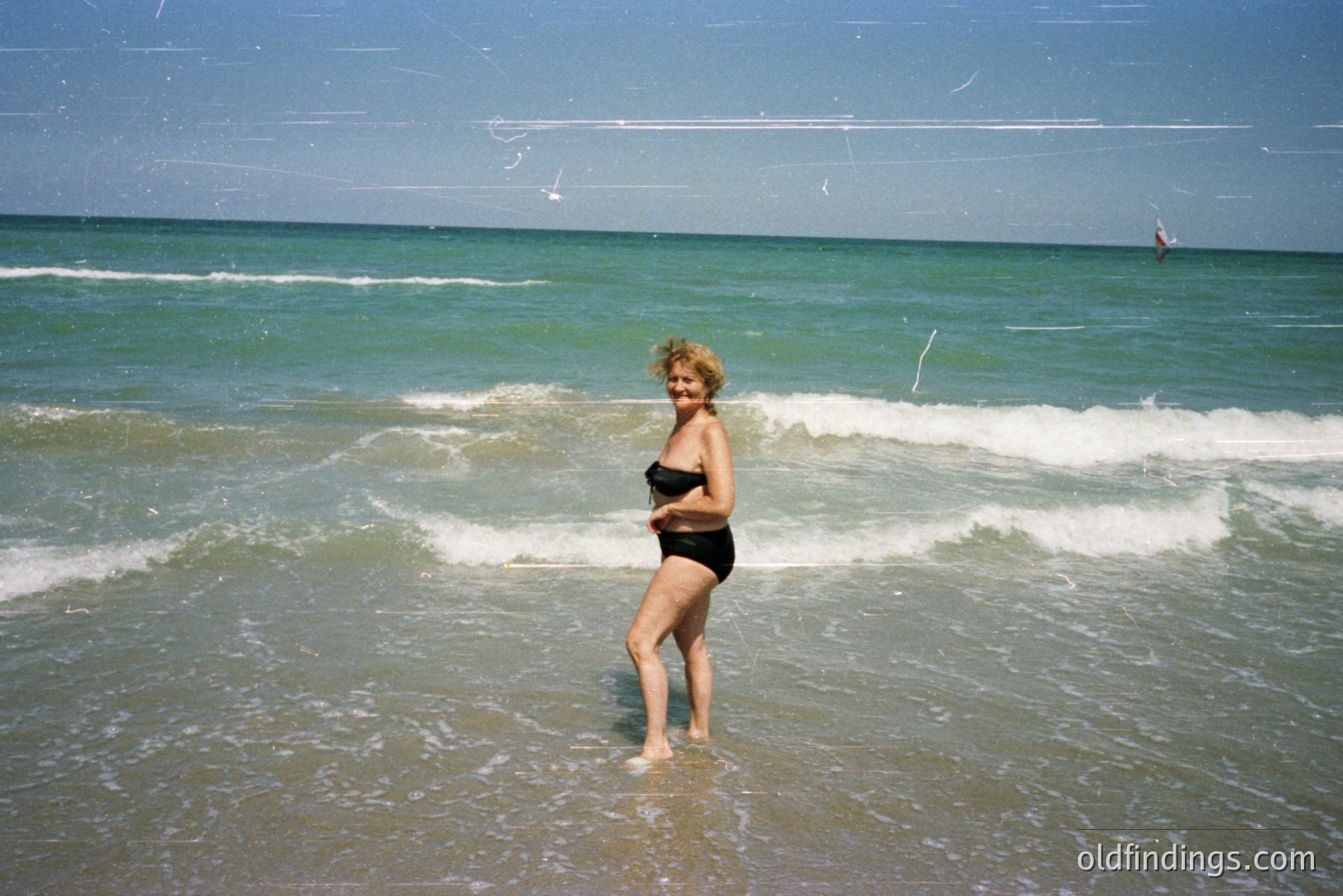 A woman stands knee-deep in the shallow waters of a beach, appearing to enjoy the waves. She wears a black bikini and has short, blonde hair. The background features a vibrant teal ocean and a clear sky. Appears to be a candid snapshot.