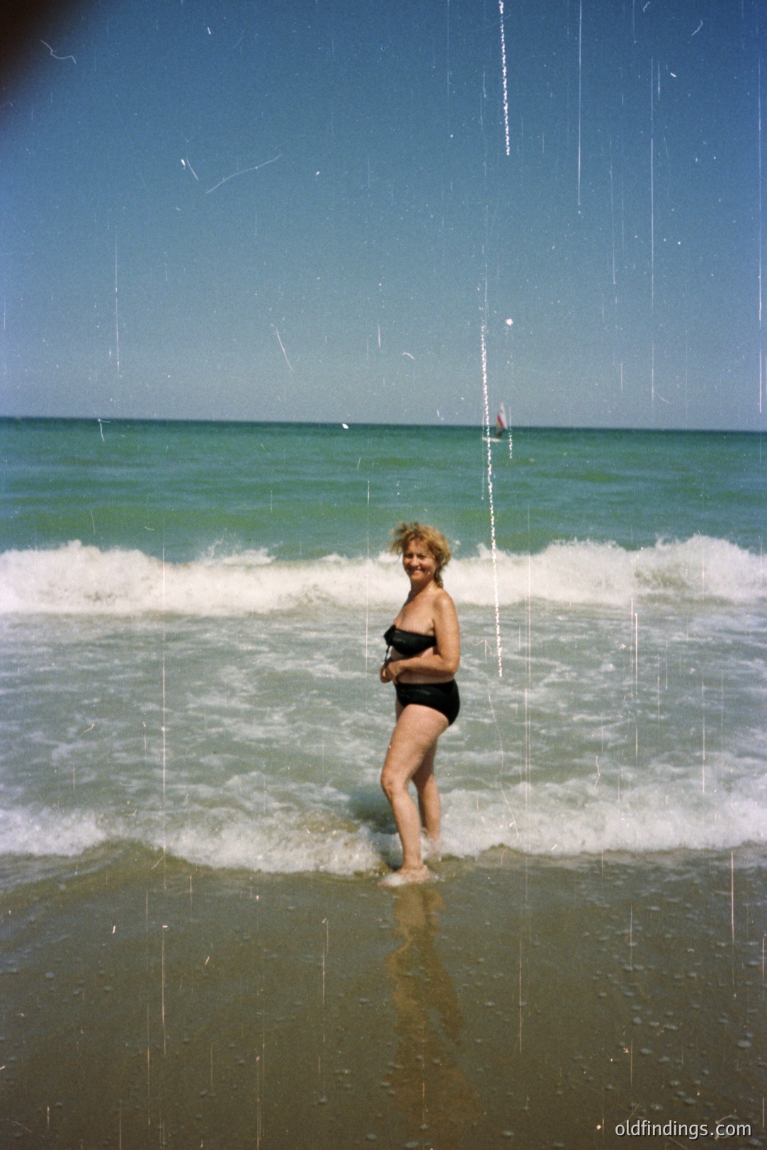 A woman stands in shallow water with waves gently lapping at her legs. She is wearing a black bikini. The bright blue sky and turquoise water suggest a warm, sunny location. Appears to be a vintage color film photograph. The sandy beach shows signs of erosion and wave action.