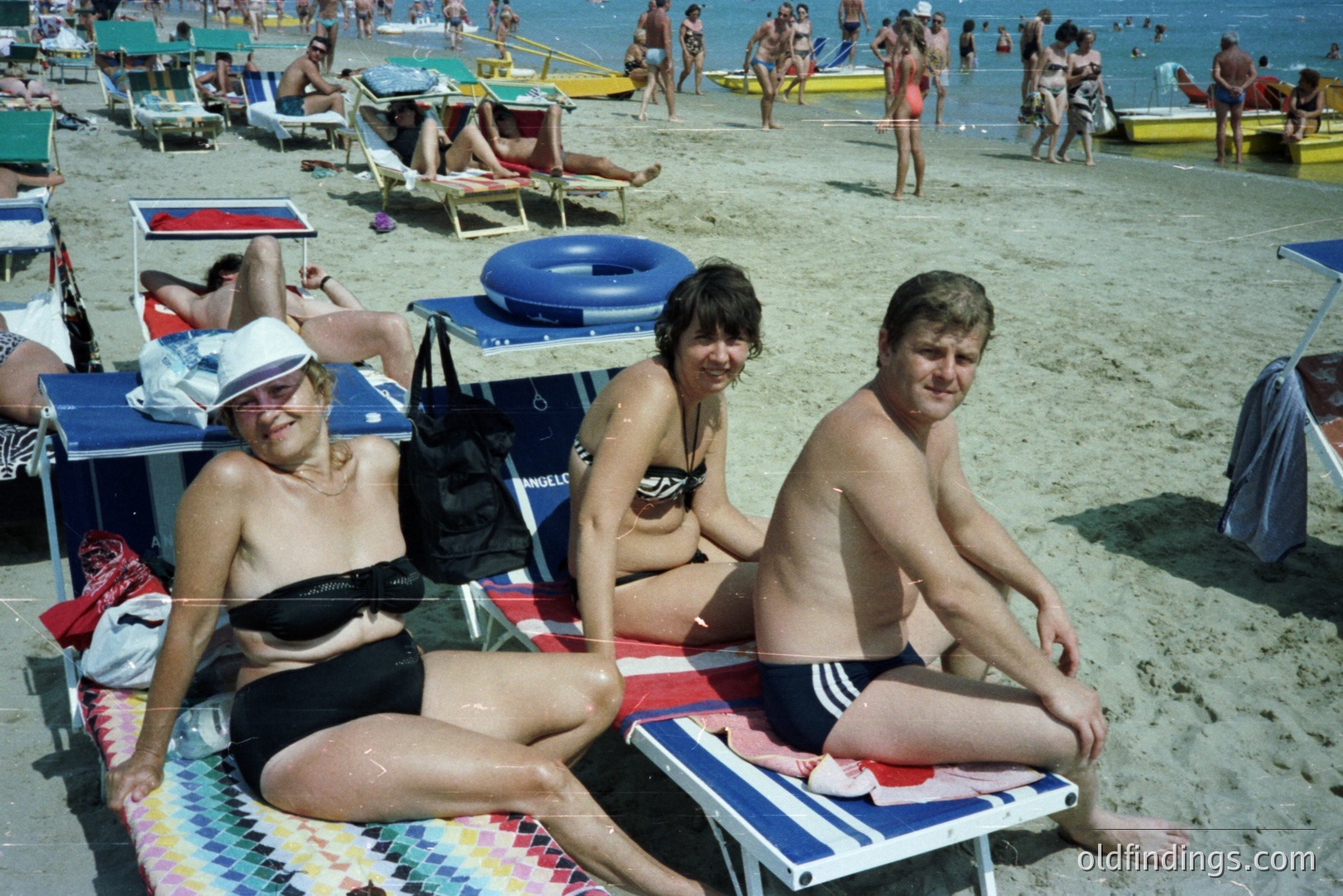 A vibrant seaside scene captures a group enjoying a sunny beach day. Three individuals, a woman in a black swimsuit and a man and woman in swimwear, sit on blue-and-white striped deck chairs, facing the camera. Background shows crowded beach with many sunbathers and boats. Likely 1970s-80s style. A nostalgic vacation snapshot.