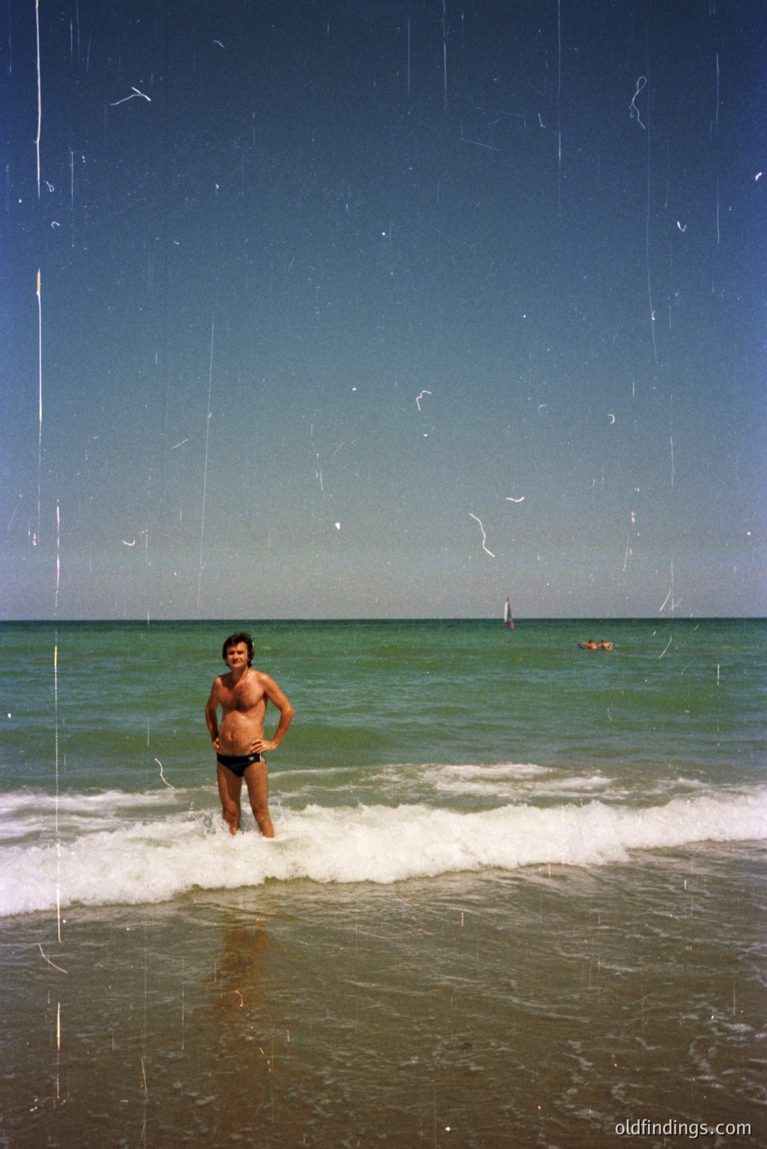 Man in patterned swim trunks stands in shallow, turquoise water along a sandy beach, facing the camera. A bright blue sky and distant swimmers complete the scene. Likely a snapshot from the 1970s, possibly a vacation photo. Visible grain and scratches indicate film photography.