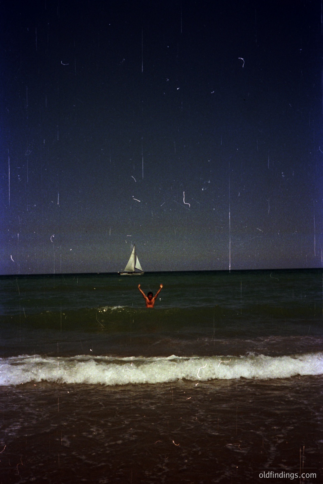 A person in an orange swimsuit extends arms as they enter waves near a sailboat under a dark sky. Grainy image suggests film photography, likely late 20th century. Evokes feelings of freedom, ocean connection. Suitable for travel, lifestyle design or vintage aesthetic projects.