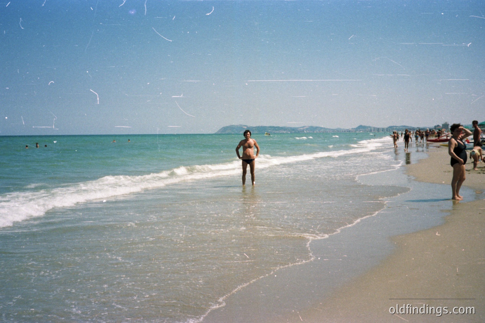 A man wades in shallow, turquoise water along a sandy beach. A woman stands nearby, both figures appearing casual. Crowd visible further down the coastline, suggesting a public beach scene. Appears to be a seaside resort, potentially Eastern Europe. Likely 1970s Kodachrome film.