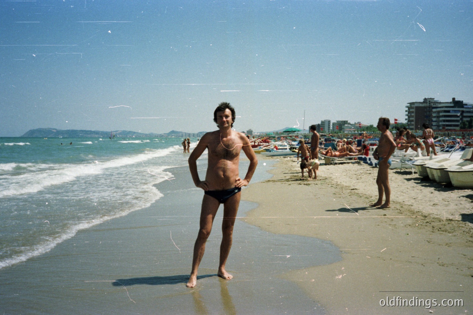 A man stands at the water’s edge on a crowded beach. He wears black swim trunks and has short, dark hair. Numerous beachgoers and colorful umbrellas populate the background. Likely a resort setting, judging by architecture. Appears to be 1970s-era snapshot.