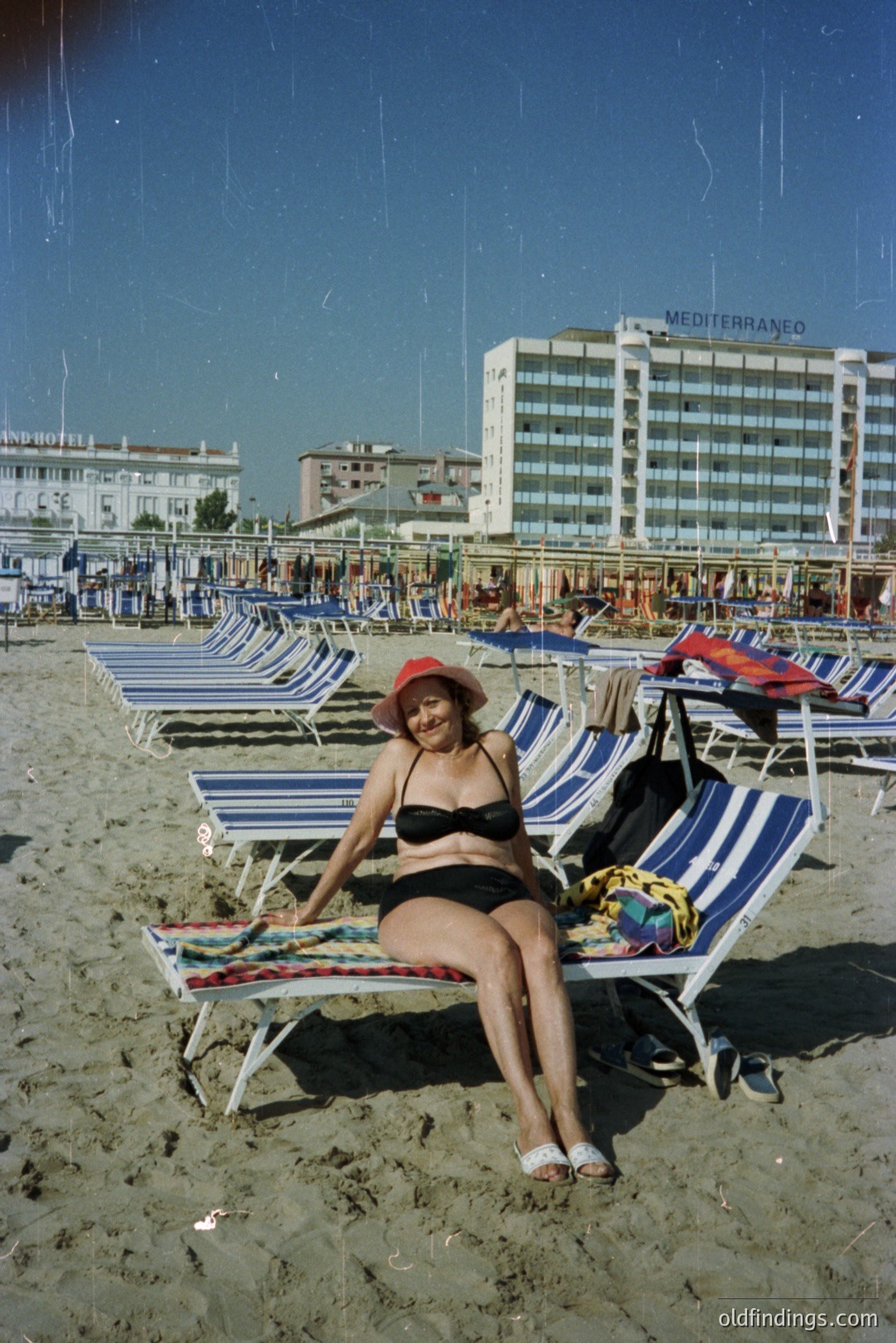 A woman sits on a blue-and-white striped beach chair, wearing a black bikini and red hat, overlooking a sandy beach and Mediterranean coastline. A large, modern hotel ("Mediterraneo") stands prominently in the background. Appears to be a 1970s travel image. The scene conveys leisure and vacation.