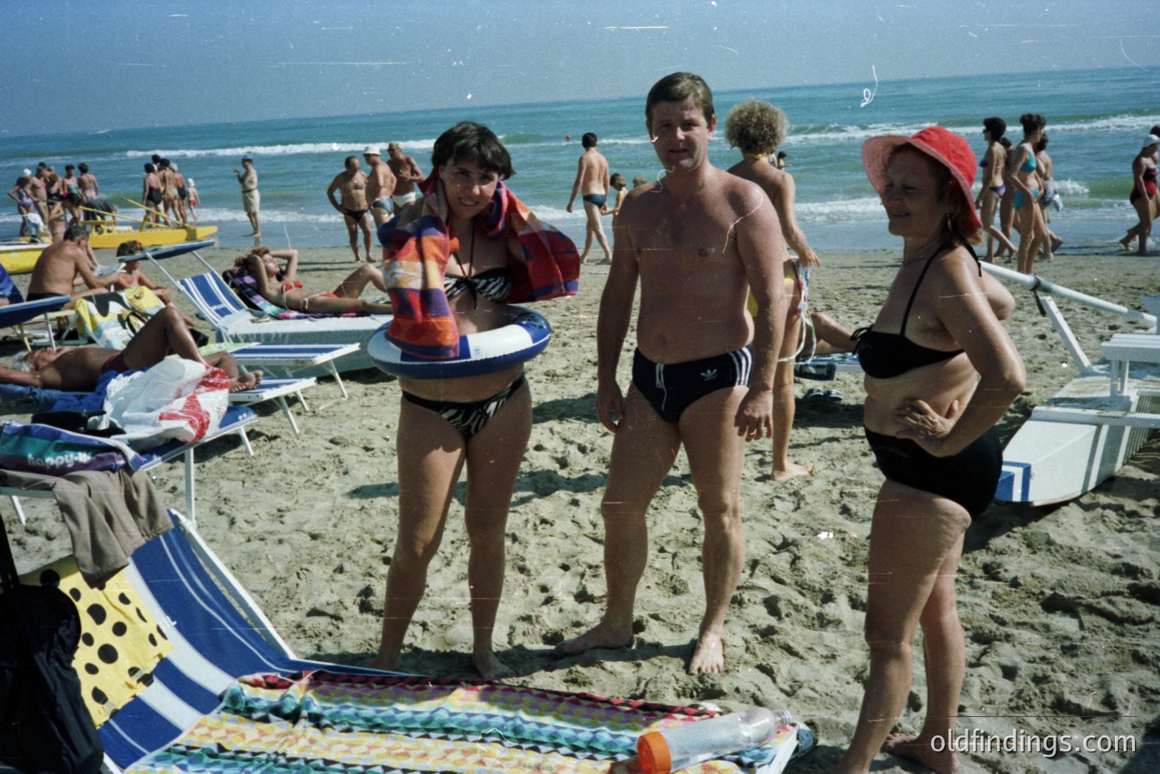 A candid snapshot of a group enjoying a crowded beach, featuring three individuals in swimwear. The man wears Adidas swim trunks; the women sport 1970s-era swimwear. Numerous beachgoers and colorful umbrellas populate the background. Likely a European coastal resort.