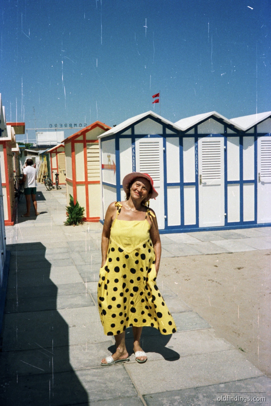 A woman in a yellow polka-dot dress and sandals poses against a row of colorful seaside cabins with red accents. The scene suggests a resort town, likely in Europe. Potential stock use for travel or fashion advertising. Vintage film grain adds character.