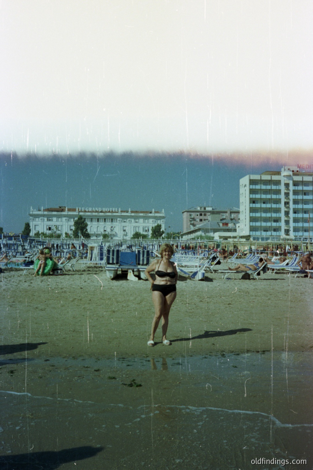 Seaside resort scene: A woman in a black bikini stands on a sandy beach with sunbathers and a hotel in the background. Likely 1970s or 80s, evidenced by the film grain & dated resort architecture. Coastal tourism imagery; potential stock photo/retro design reference.