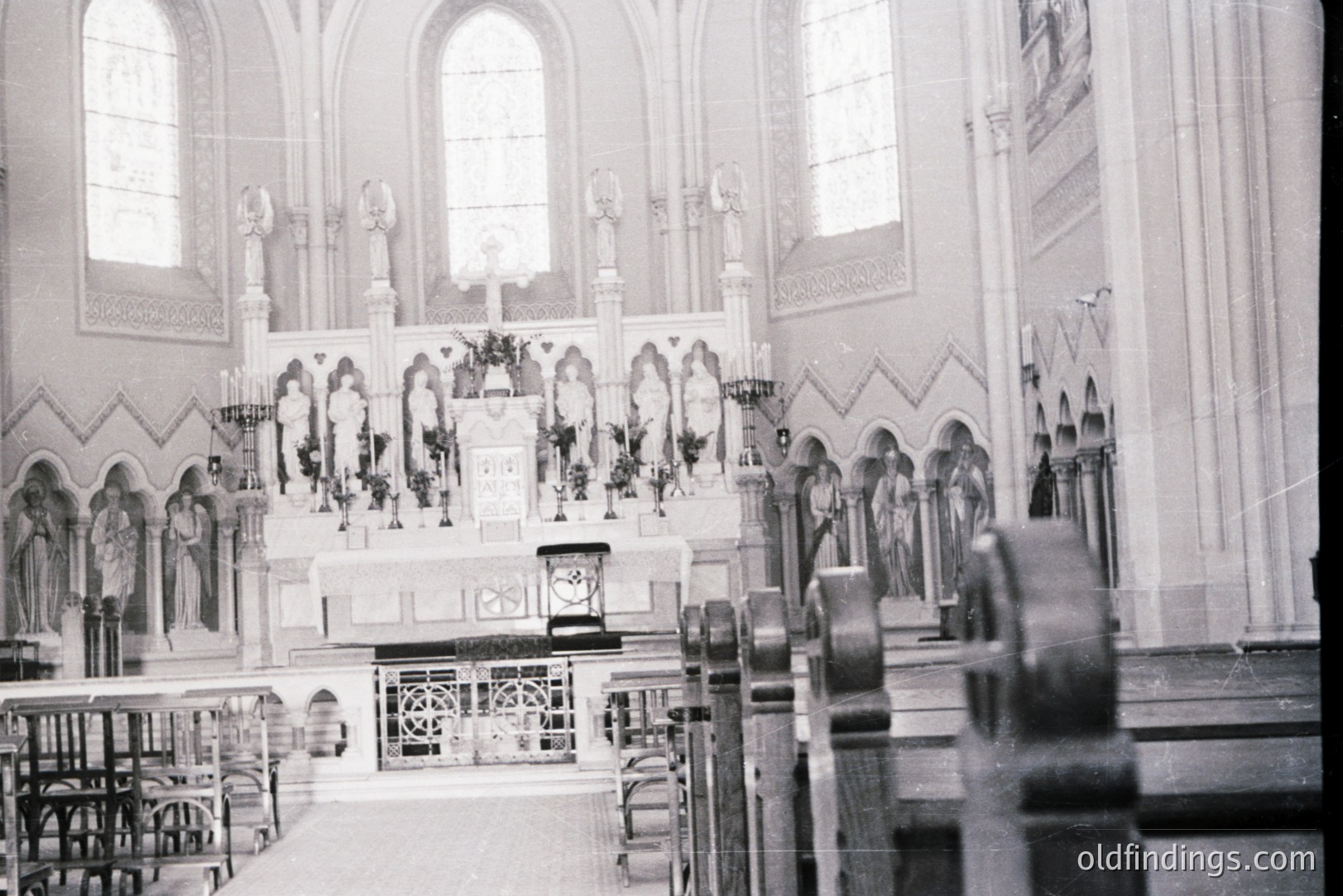 Interior view of a Catholic church, showcasing an ornate altar with statues, floral arrangements, and a crucifix. The architecture features pointed arches and stained glass windows. Likely from the early to mid-20th century, possibly a European location. Valuable for architectural reference and religious studies.