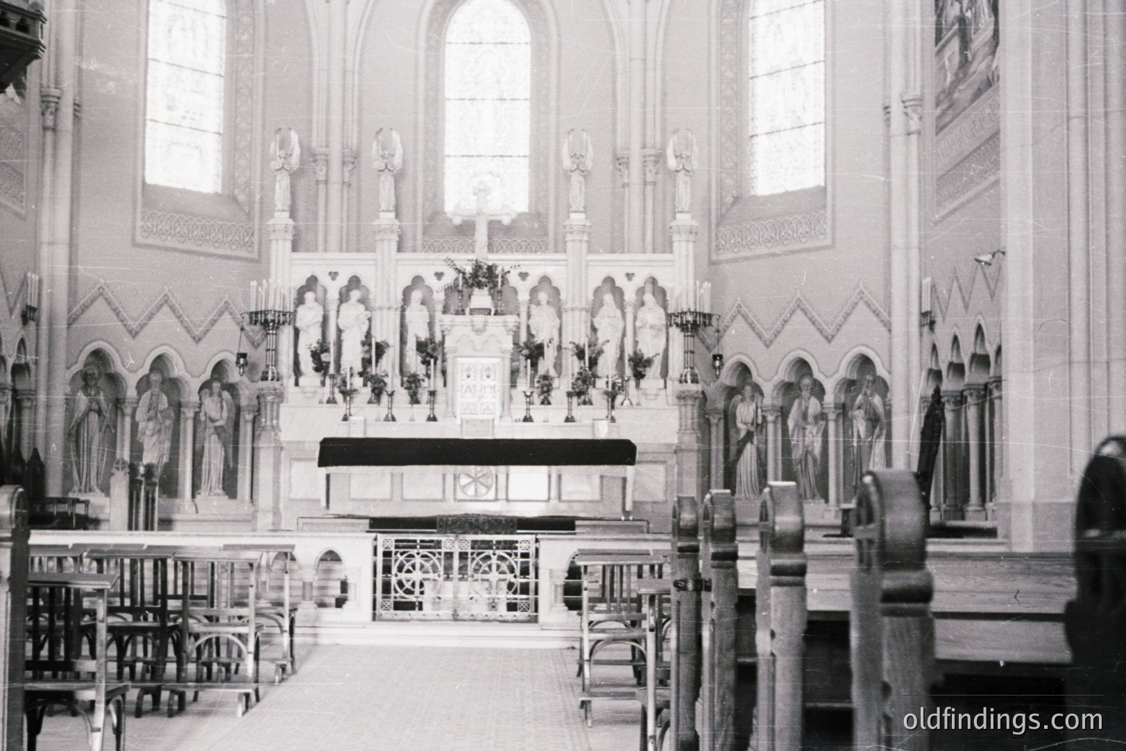 Ornate, black and white photo of a Catholic church interior. Focus on the altar, adorned with floral arrangements & statues. Features include pointed arches, decorative woodwork, and rows of pews. Likely mid-20th century, judging by the photographic style & architectural detailing.
