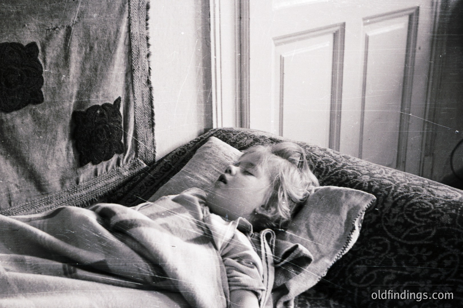 A young child sleeps peacefully, bundled in a blanket, within a bedroom. Detailed wallpaper and a dark floral throw provide rich texture. The photograph, likely from the 1930s or 40s, evokes a sense of domestic tranquility and childhood innocence. A decorative wall hanging is partially visible.