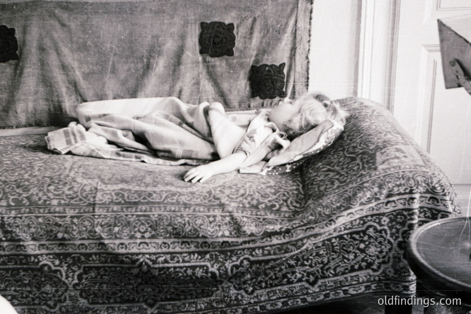 A child rests on an ornate, patterned sofa beneath a decorative textile. The scene suggests a domestic interior, possibly mid-20th century based on the photographic style and decor. A table leg is visible in the right foreground. Likely a stock photography reference or historical document.