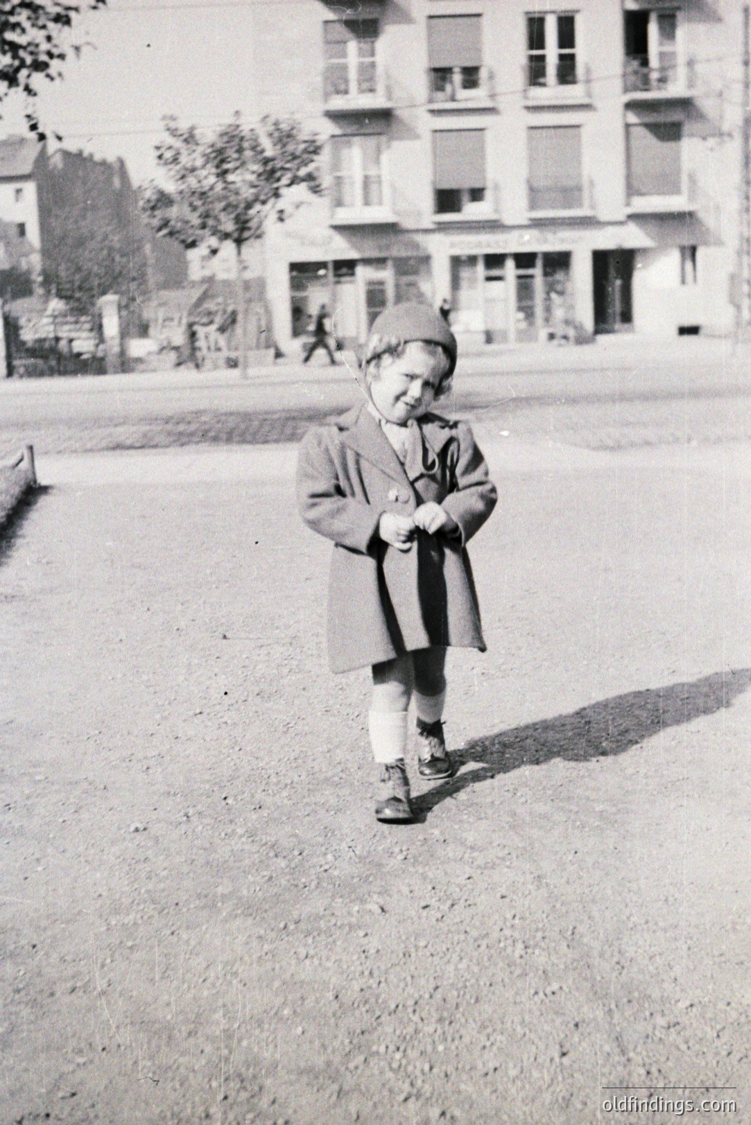 A young child stands outdoors, wearing a warm coat, patterned stockings, and sturdy shoes. A multi-story building with large windows forms the backdrop. Likely 1950s-1970s, capturing a candid moment of childhood. The grainy texture suggests a vintage print. Potential for historical family archives or design inspiration.