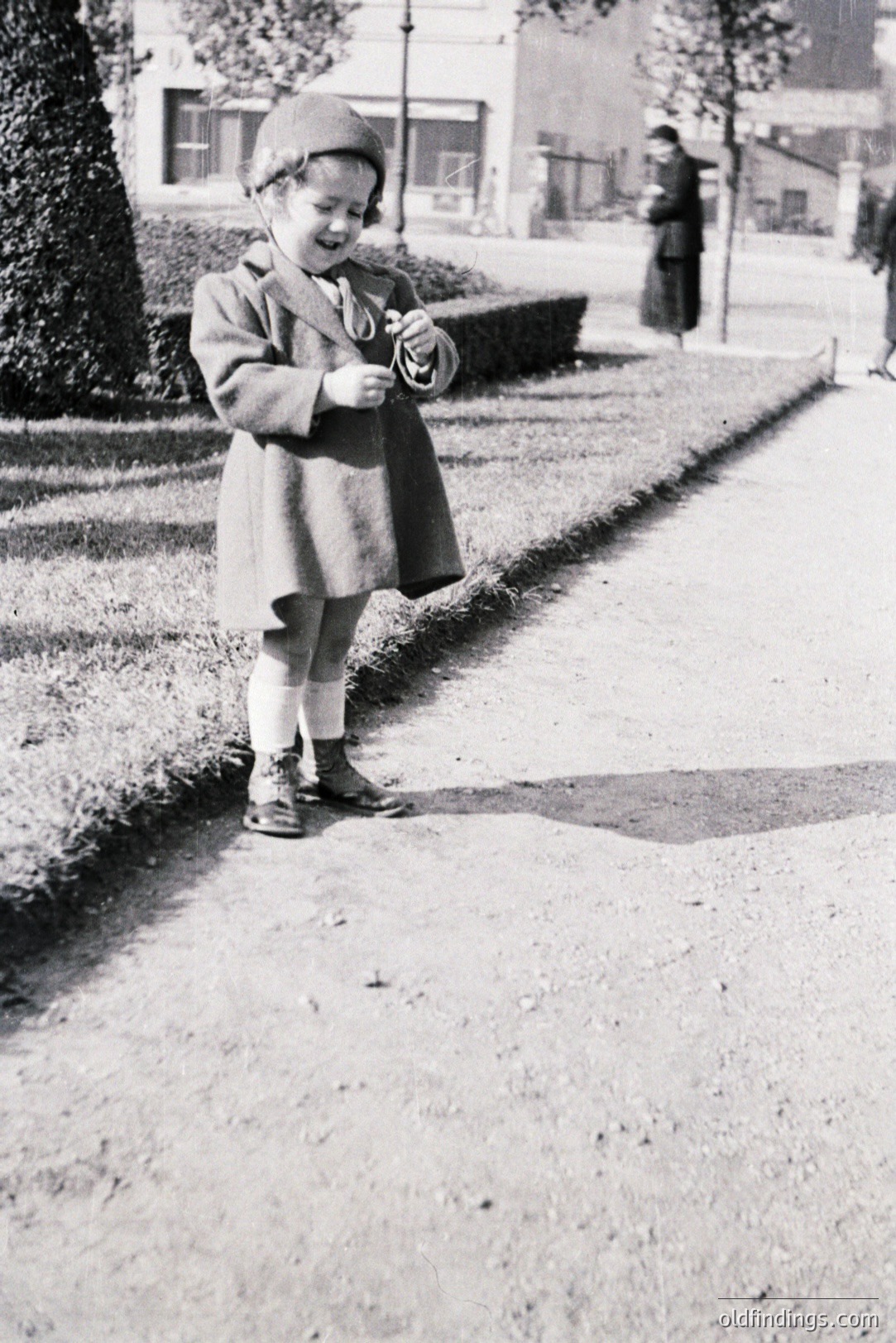 A young child, bundled in a wool coat and hat, stands near a manicured hedge. White stockings and sturdy shoes complete the outfit. A woman in a long dress is visible in the background. Possibly a portrait taken in a European park. Likely 1940s-1960s.