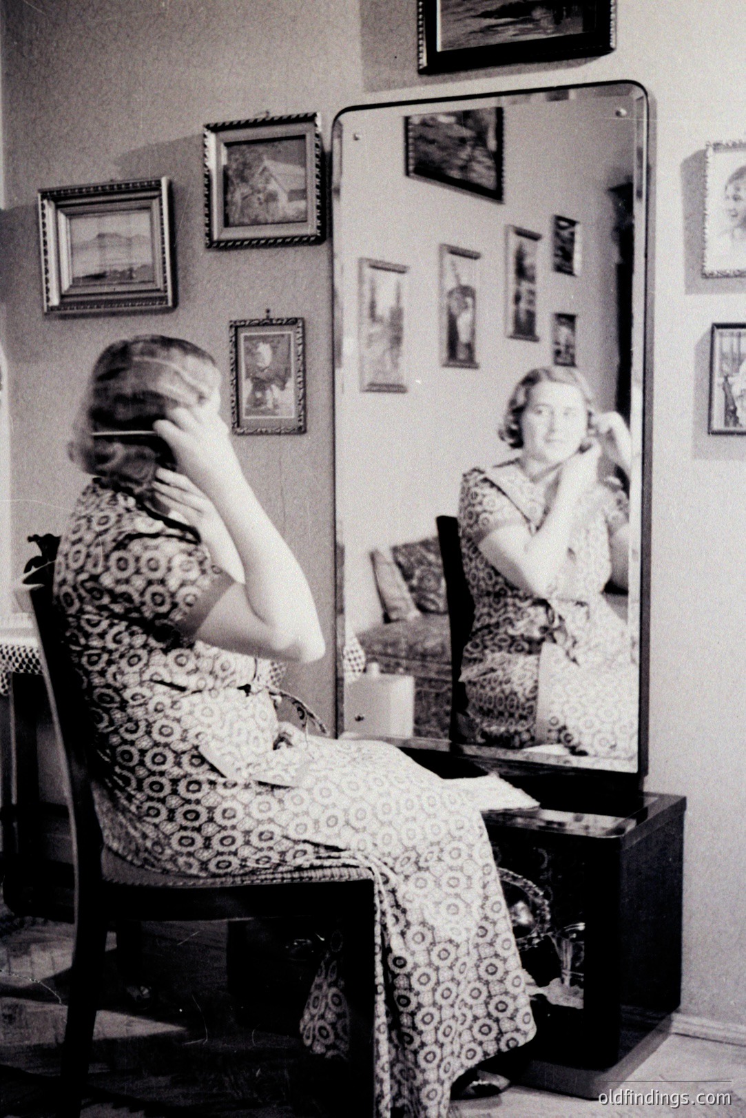 A young woman sits before an ornate mirror, seemingly fastening jewelry. The room features a decorative dresser and multiple framed pictures on the wall. Likely a domestic interior, style evokes the 1940s-1960s. Fine art reference and vintage style imagery.