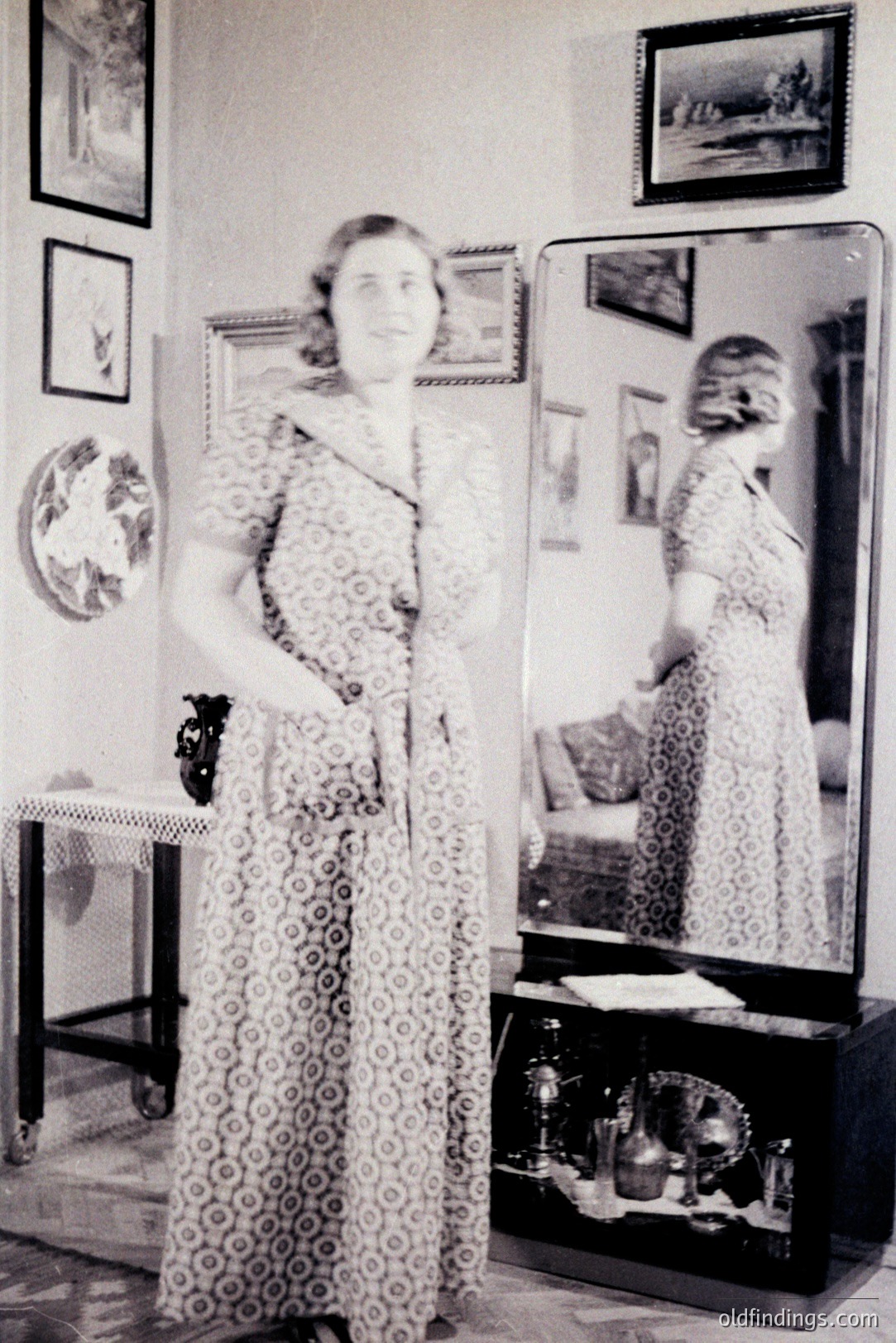 Full-length portrait of a woman in a patterned dress standing before a tall, ornate mirror. Interior setting with decorative side table, trophies, and framed artwork. Likely 1930s-1950s, judging from dress style and photographic aesthetic. Reflective surface adds depth.