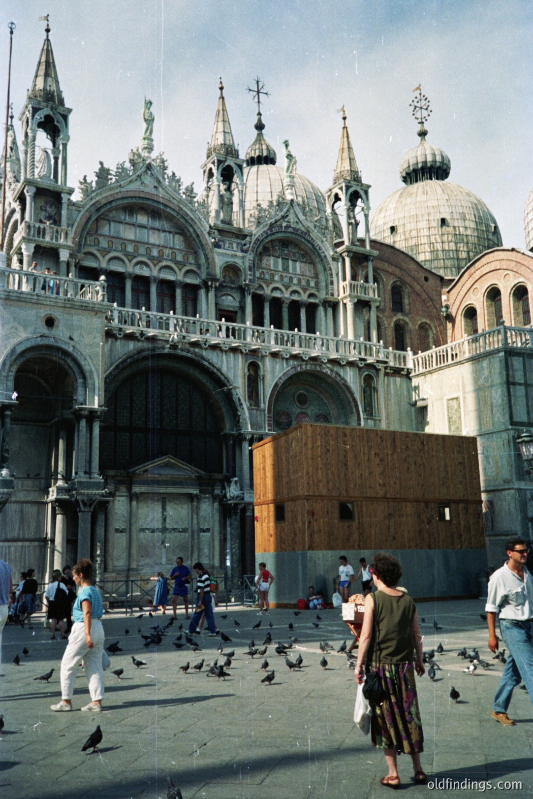St. Mark's Basilica, Venice, with visible scaffolding indicating restoration work. A crowd of tourists, pigeons, and a few local residents populate the piazza. The architectural style is Venetian Gothic, characterized by elaborate domes and arches. Likely captured in the 1980s-1990s based on attire.