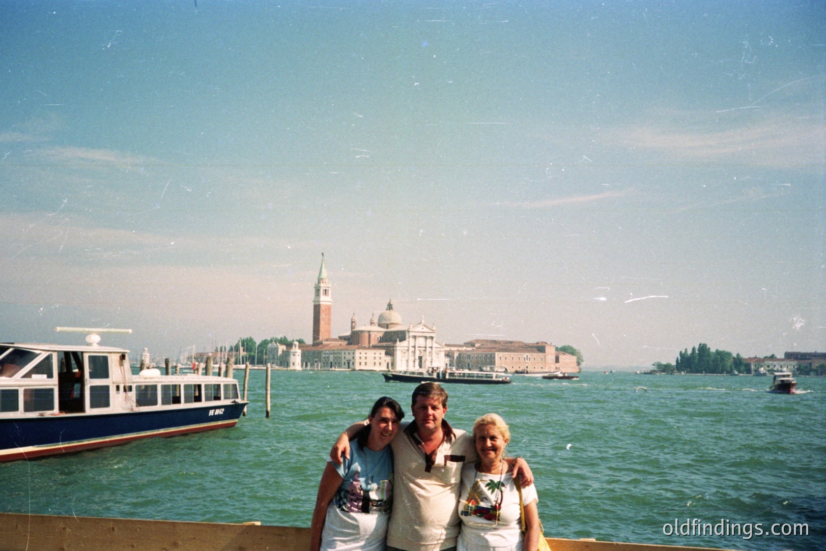 A family of three poses for a photo with San Giorgio Maggiore island and Venice in the background. The subjects, likely tourists, are casually dressed. A boat is visible to the left. Likely taken in the 1990s, judging by the photographic style and attire.