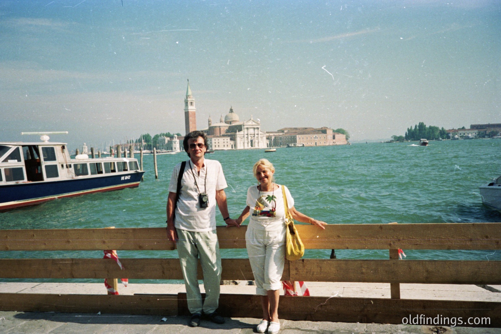A couple stands near a weathered railing, holding hands, with San Giorgio Maggiore island visible across the Venetian lagoon. The man wears a camera around his neck; the woman sports a floral shirt. Appears to be a tourist snapshot, c. 1990s.