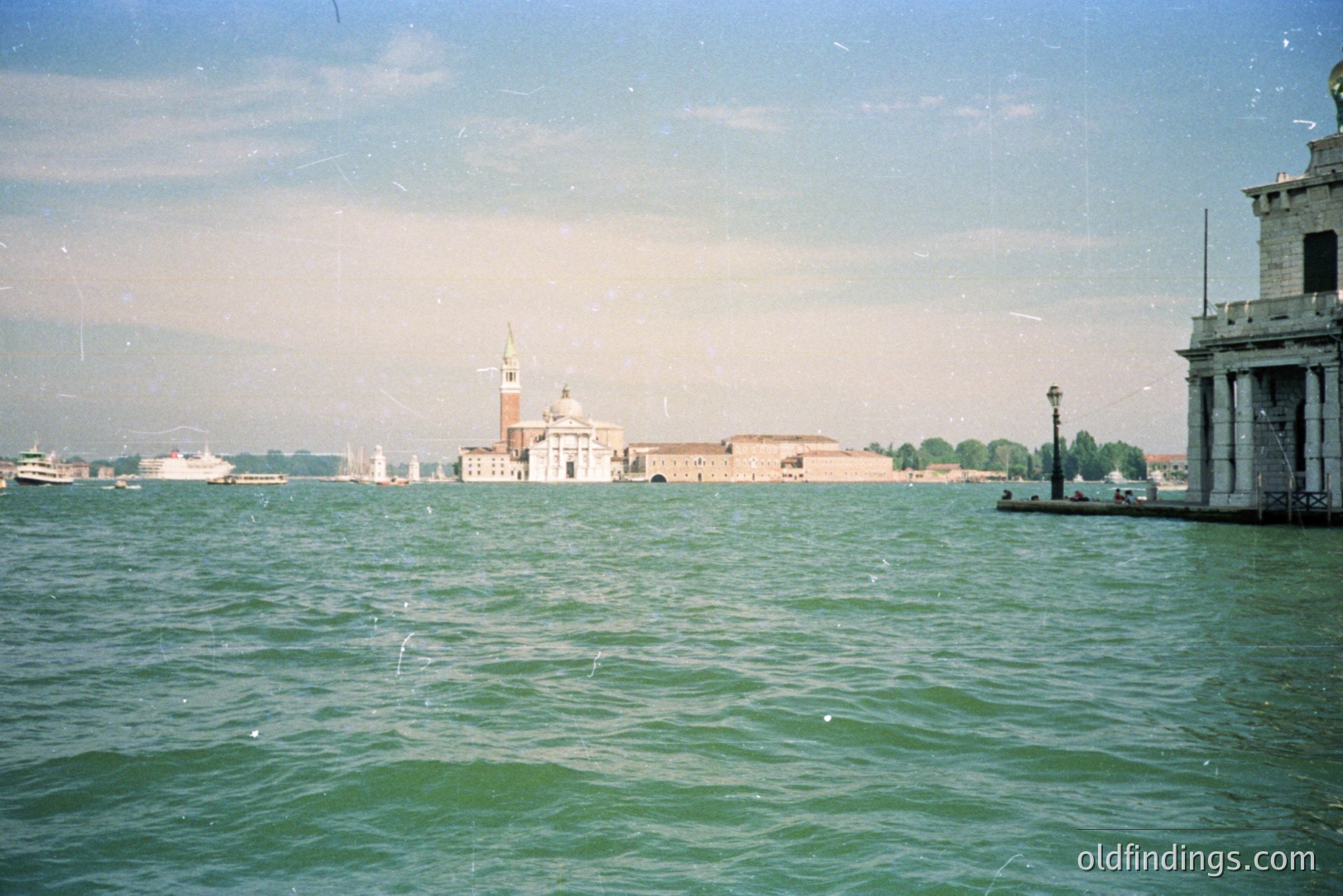 A long-shot view of Venice, Italy, showcasing San Giorgio Maggiore island & its bell tower across the lagoon. A stone pier occupies the foreground, with distant sailboats visible. Likely a tourist image, capturing a classic Venetian scene.