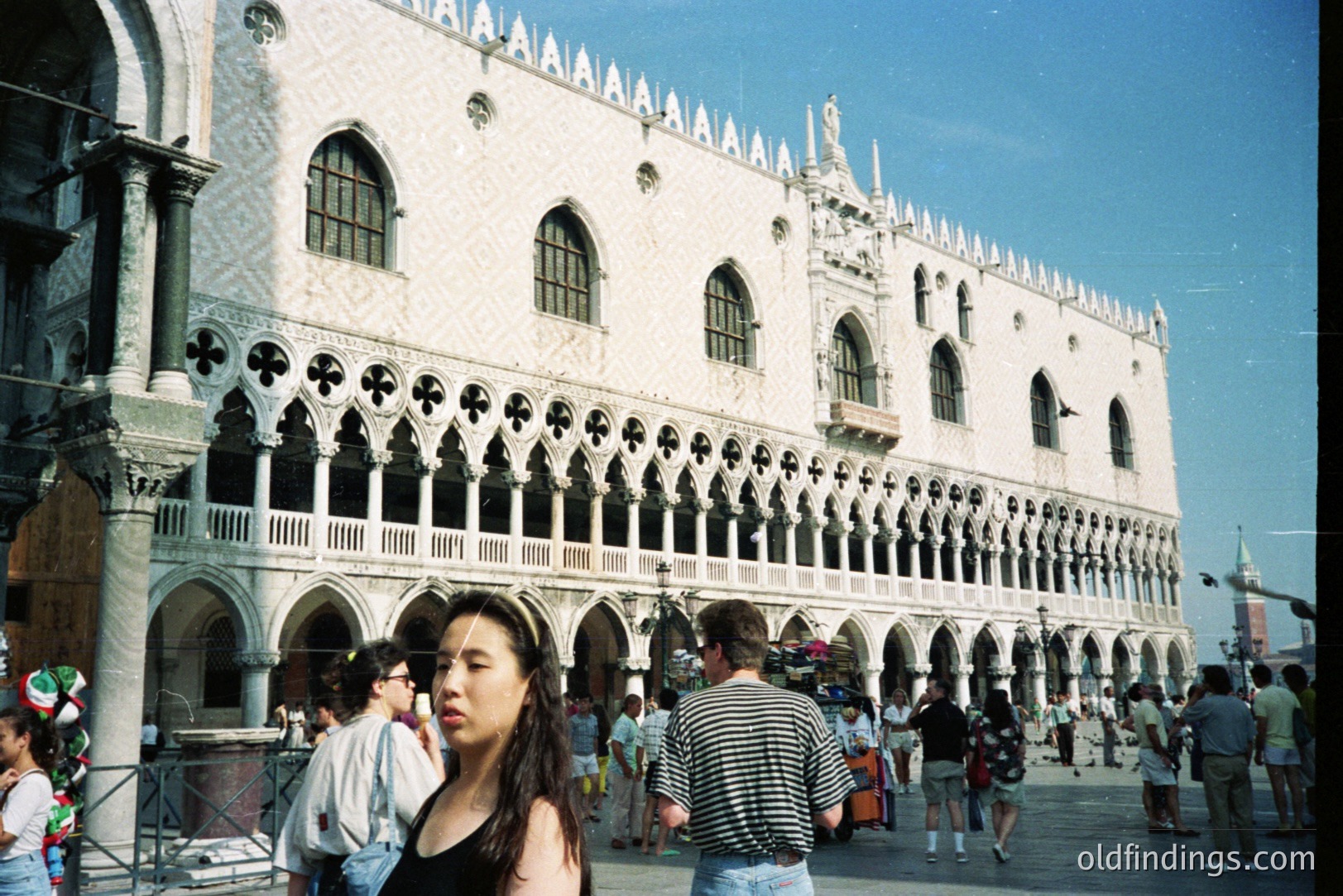 An exterior view of the Doge's Palace in Venice, Italy, featuring its iconic arcaded facade. A young woman stands in the foreground, partially obscuring the bustling Piazza San Marco. The photograph’s color palette suggests a 1990s snapshot, capturing a moment of tourism and architectural grandeur.