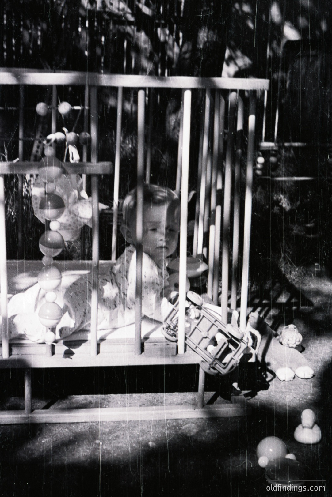 A young child sits within a wooden crib, surrounded by toys and balloons. The photograph’s composition is centered on the child, capturing a moment of childhood play. Likely a candid snapshot, the image evokes a sense of nostalgia. Estimated timeframe: 1950s-1970s.