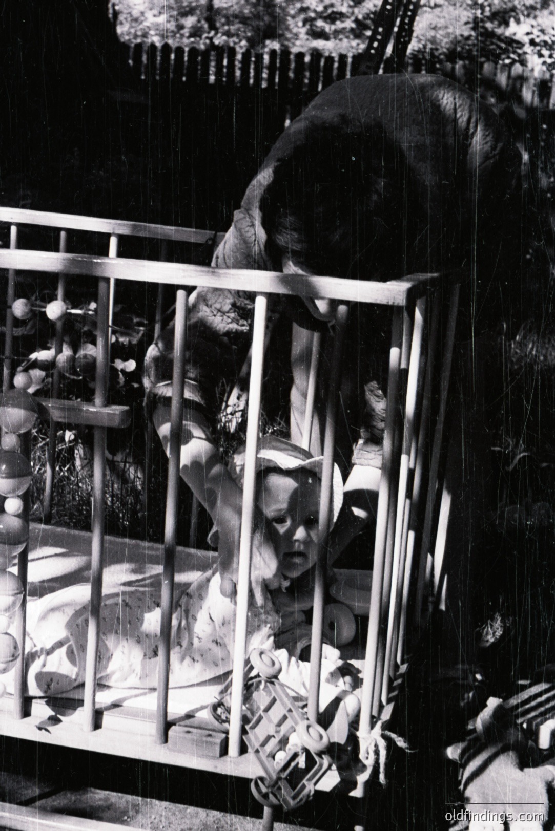 A young child sits within a wooden crib, surrounded by toys, appearing to observe an elephant looming behind the structure. Possibly a zoo exhibit or private menagerie. The photograph's grainy quality suggests a snapshot from the mid-20th century. A poignant, somewhat surreal moment captured.