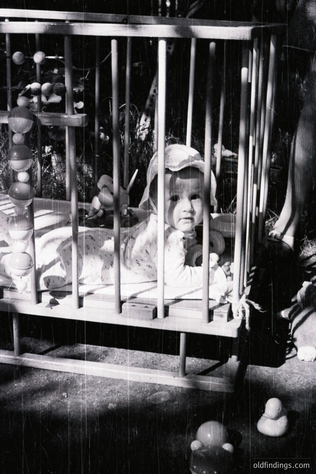 Infant lies within a vintage wooden crib, gazing directly at the camera. A mobile with wooden beads hangs overhead. Likely taken in a domestic setting, possibly a garden. The photograph’s style suggests a mid-century aesthetic, circa 1950s-1970s. Valuable for nostalgic design references.