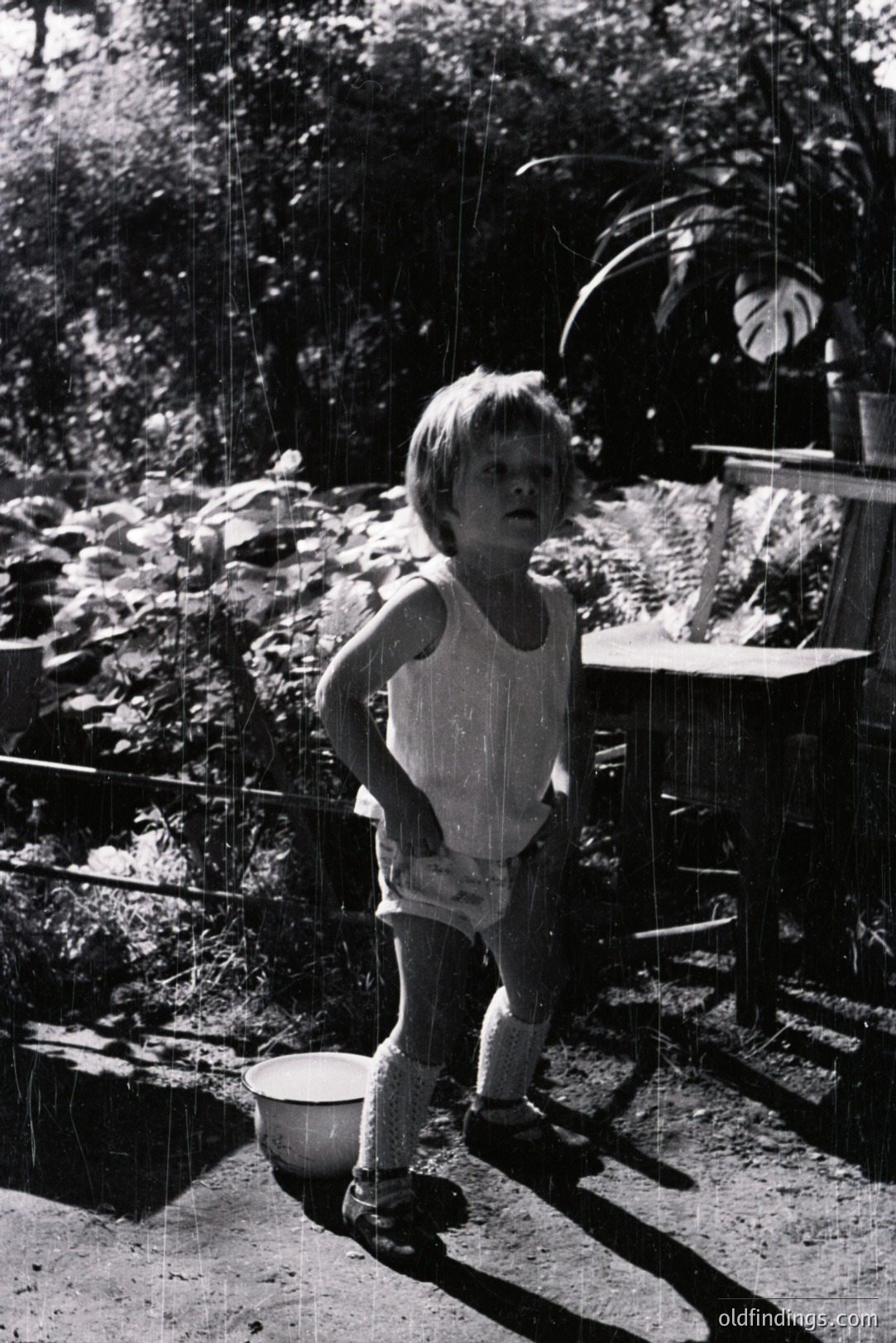 A young child stands playfully in a light rain, captured in a candid moment. The subject wears a tank top and knee-high socks, their posture suggesting youthful energy. A metal bucket sits nearby amidst lush foliage and a weathered table. Likely mid-20th century, the grainy texture and composition are characteristic of period snapshots.
