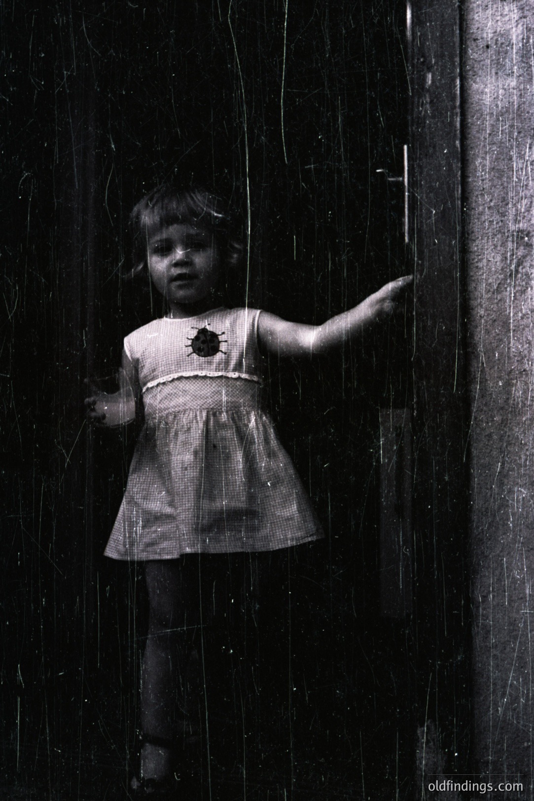 A young girl in a patterned dress stands peering out from behind a heavily scratched window, arm extended towards the door handle. The stark contrast evokes a sense of curiosity and confinement. Likely a candid moment, possibly 1950s-1970s, with potential for stock or archival use.