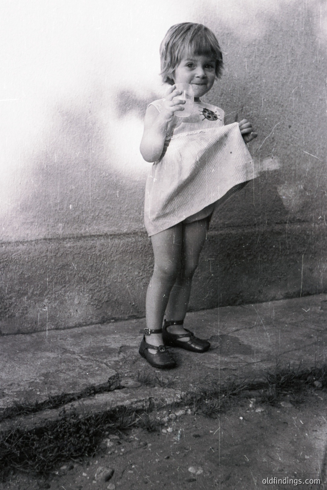 A young girl, circa 1960s-1970s, stands near a weathered stone wall, playfully holding up the hem of a sleeveless, lace-trimmed dress. She wears Mary Jane shoes with ankle straps. The grainy image exhibits signs of age, with surface scratches. Likely a snapshot from family archives.