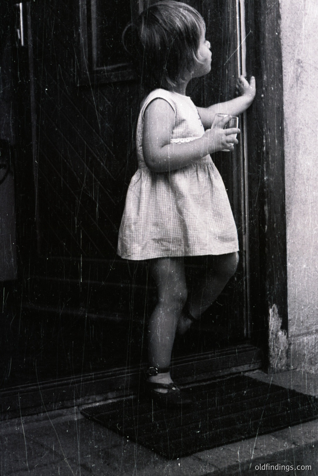 A young child in a short dress and sandals stands on a welcome mat, gently touching a door frame. The image, likely from the 1960s or 70s, exhibits signs of age, including visible grain and surface scratches. A simple, domestic scene.