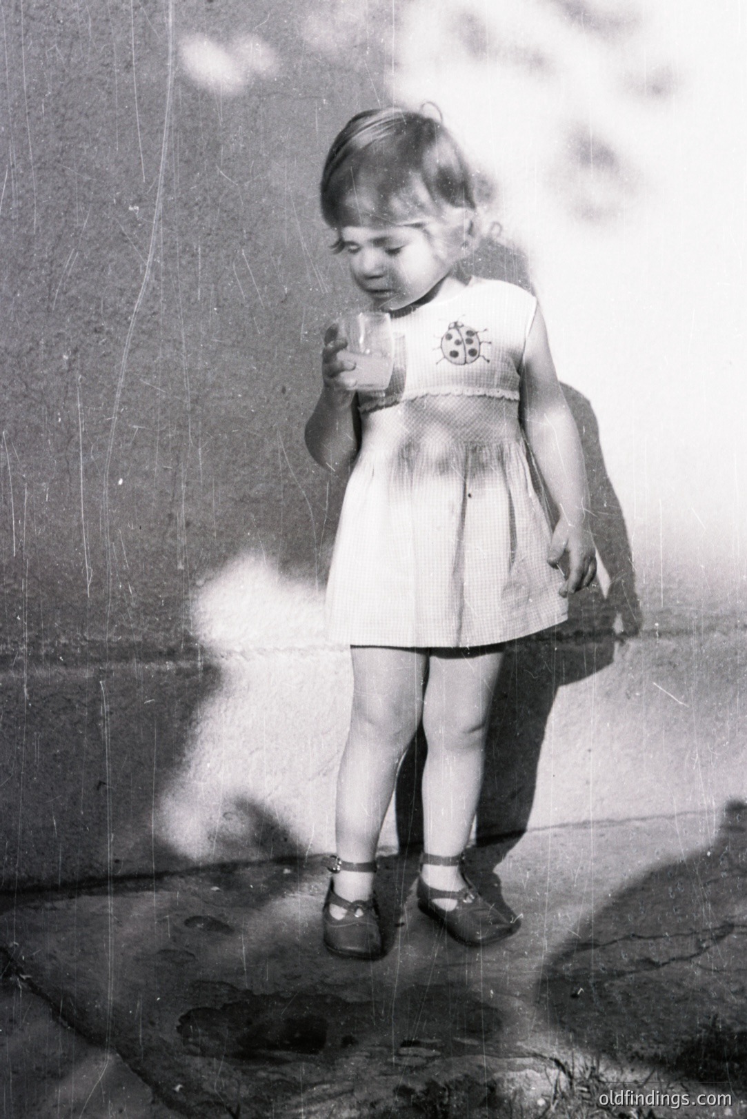 A young girl stands holding a glass, her shadow cast long behind her. She wears a dress with ruffled detailing and sandals with ankle straps, typical of mid-20th century children's wear. The image exhibits wear consistent with age and storage. Likely 1950s-1970s.