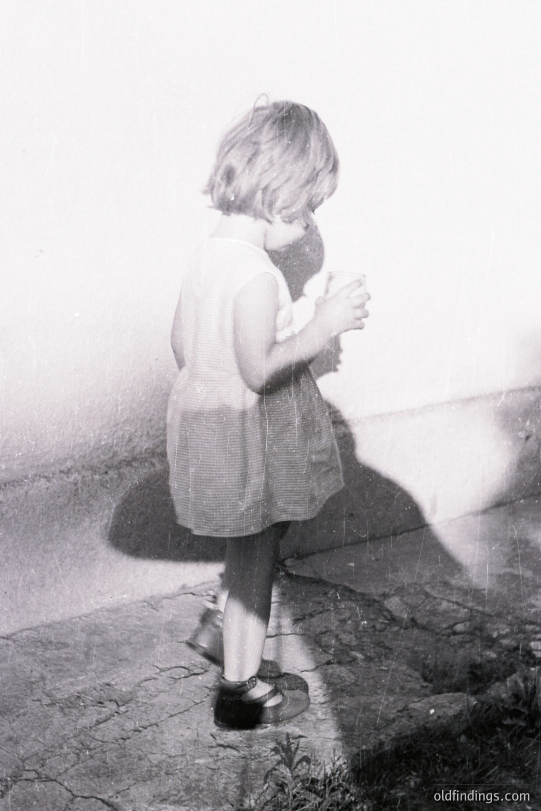 A young girl in a simple dress and Mary Jane shoes examines something held in her hands against a textured wall. The photo's grainy quality and style suggests a candid moment, likely from the 1960s or 1970s. The backdrop features cracked pavement and sparse vegetation. Simple, nostalgic domestic scene.