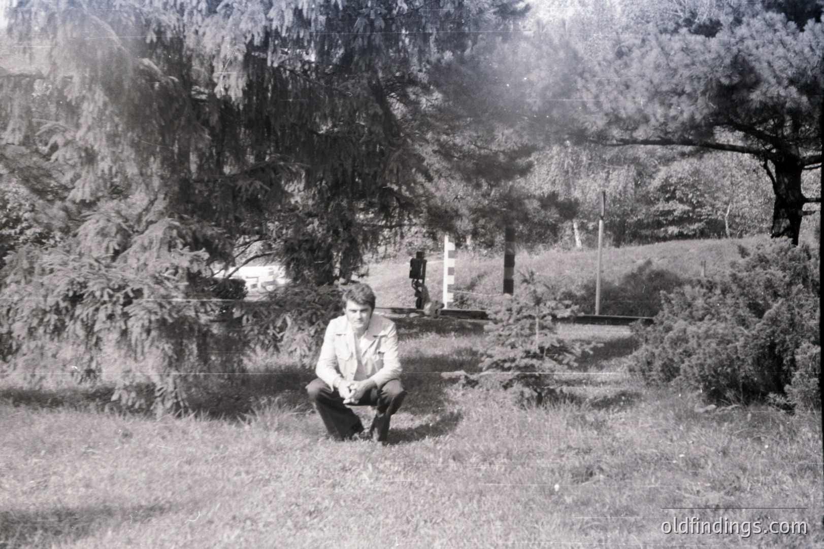 A young man, dressed in a light-colored collared shirt and dark trousers, squats within a grassy area, flanked by dense foliage. Appears to be a landscaped park or garden setting. Possibly a snapshot from the 1960s or 1970s, showing mid-century fashion. Amateur photography style.