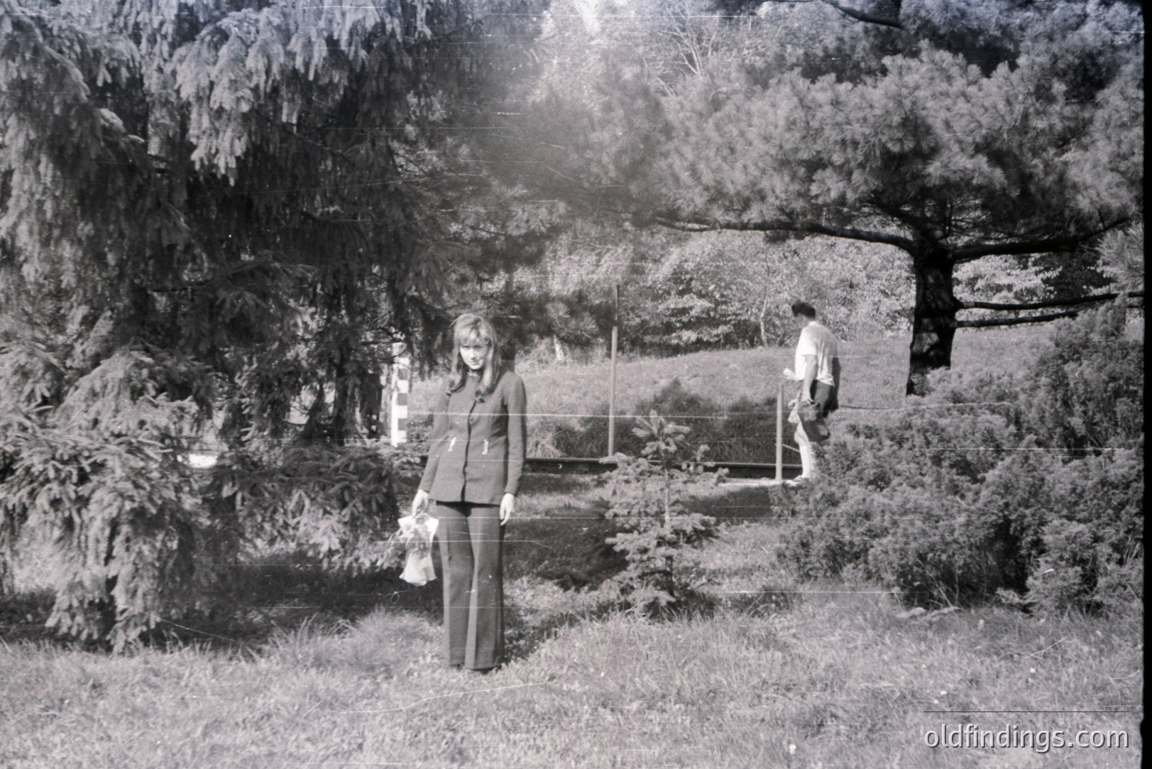 Monochrome image depicts a woman in a wide-leg trouser suit, holding a shopping bag, standing on grassy terrain with dense shrubbery & trees. A figure walks away in background. Likely a 1970s fashion or lifestyle scene. Possible park or garden setting. Archive/vintage aesthetic.
