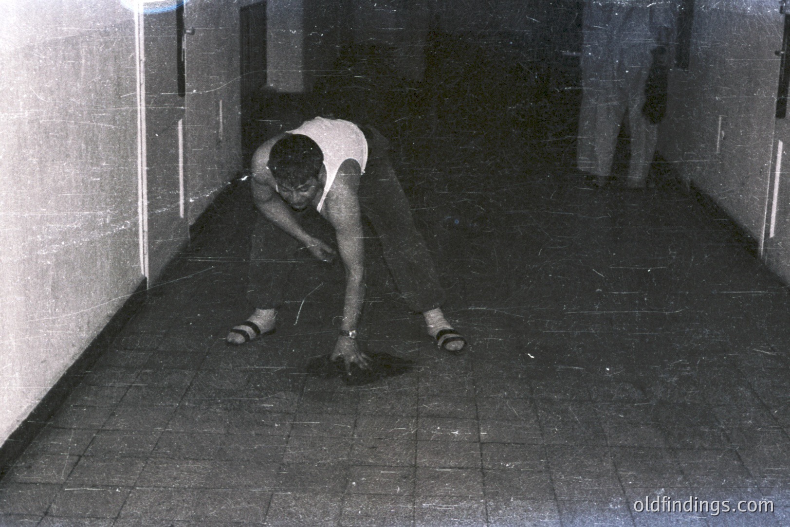 A man in a sleeveless shirt and sandals appears to be gesturing emphatically on a tiled floor within a narrow hallway or corridor. The scene is dimly lit, with several closed doorways lining the walls. Visible grain suggests film photography. Likely 1970s-era architectural style. Stock potential for design/mood.