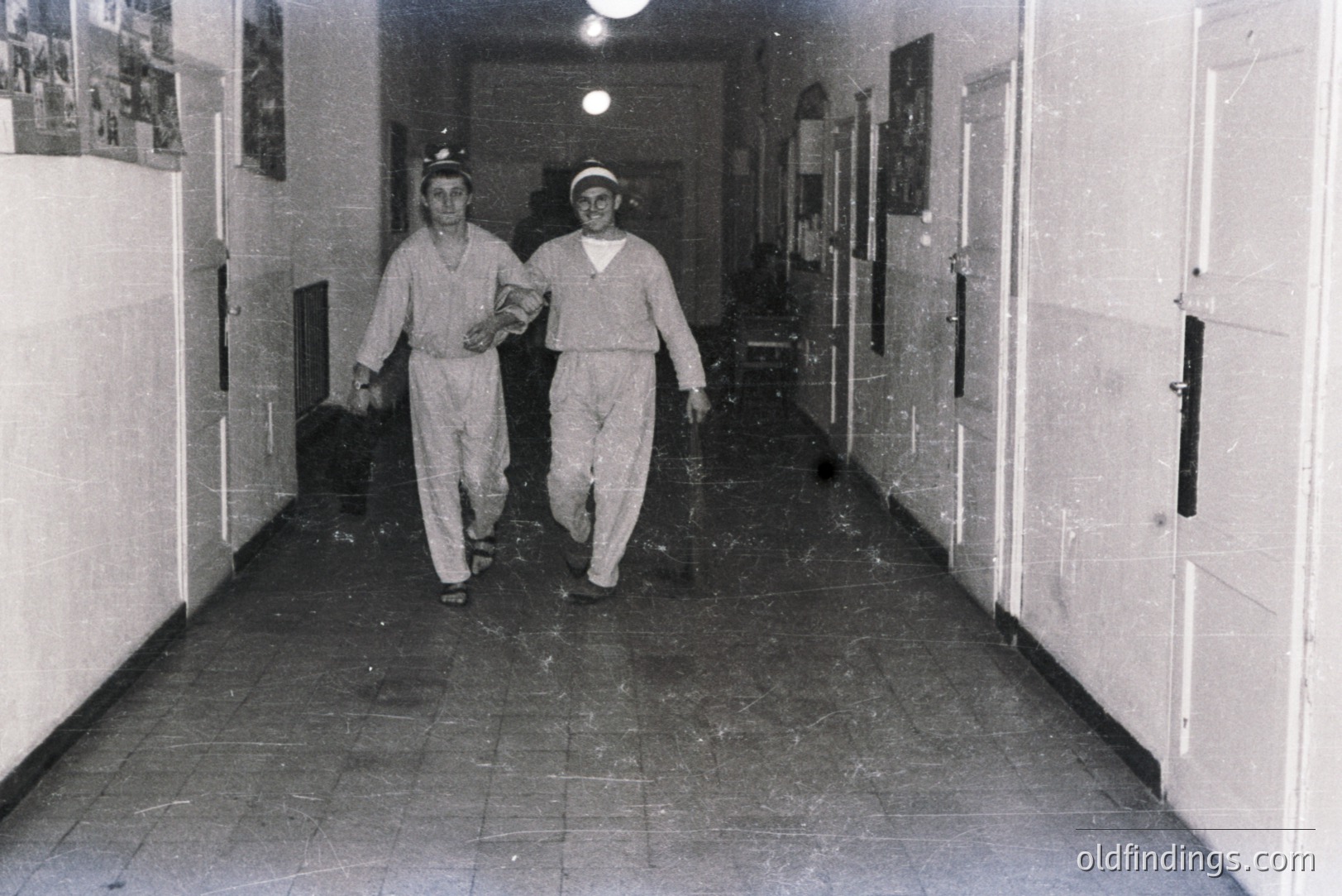 Two men in uniform and headgear walk down a tiled hallway. The scene appears to be an institutional setting, possibly a hospital or military facility. Interior architecture features closed doors lining both sides of the corridor. Dust and grain evident on film. Likely 1950s-1970s.