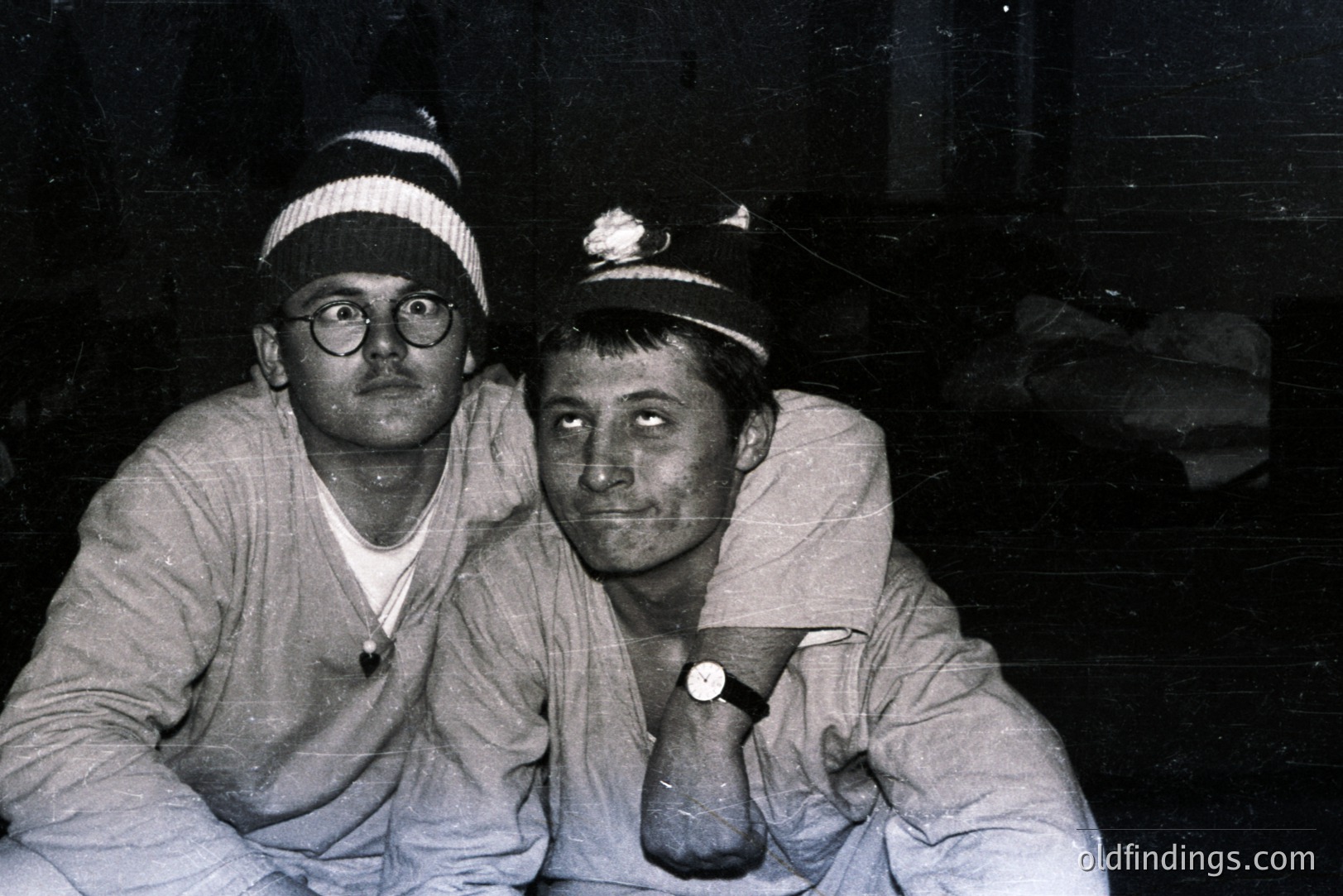 Two men wearing knit hats sit close together; one with glasses and a white t-shirt, the other with a visible wristwatch. The image exhibits signs of age, like graininess, and looks like a candid or informal portrait. Likely 1960s-1970s, possibly depicting a rural or working-class lifestyle.