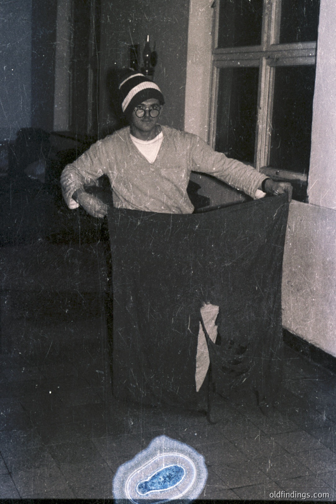 A man in a dark sweater and spectacles poses with a large, dark fabric draped over his body, seemingly held up by his arms. Visible architectural details suggest an interior space with a window. The photo exhibits signs of age, including dust and scratches. Likely mid-20th century, style evokes a vintage portrait.