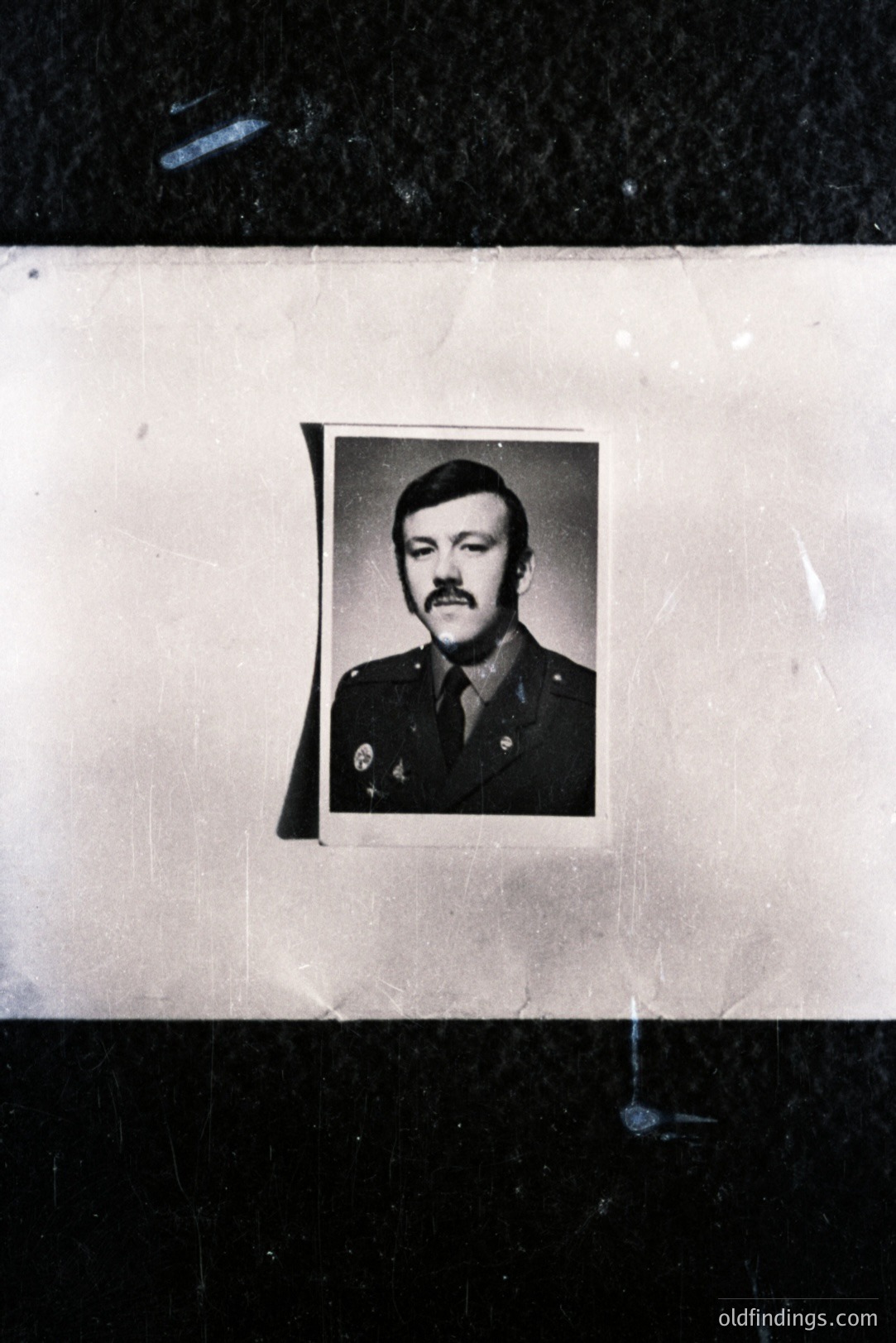 Formal portrait of a man in military uniform, featuring a prominent mustache and neatly styled hair. Likely a service or personnel photo, displaying rank insignia on the shoulder. The photo appears aged, mounted on a textured surface, exhibiting signs of wear. Appears to date from the 1970s.