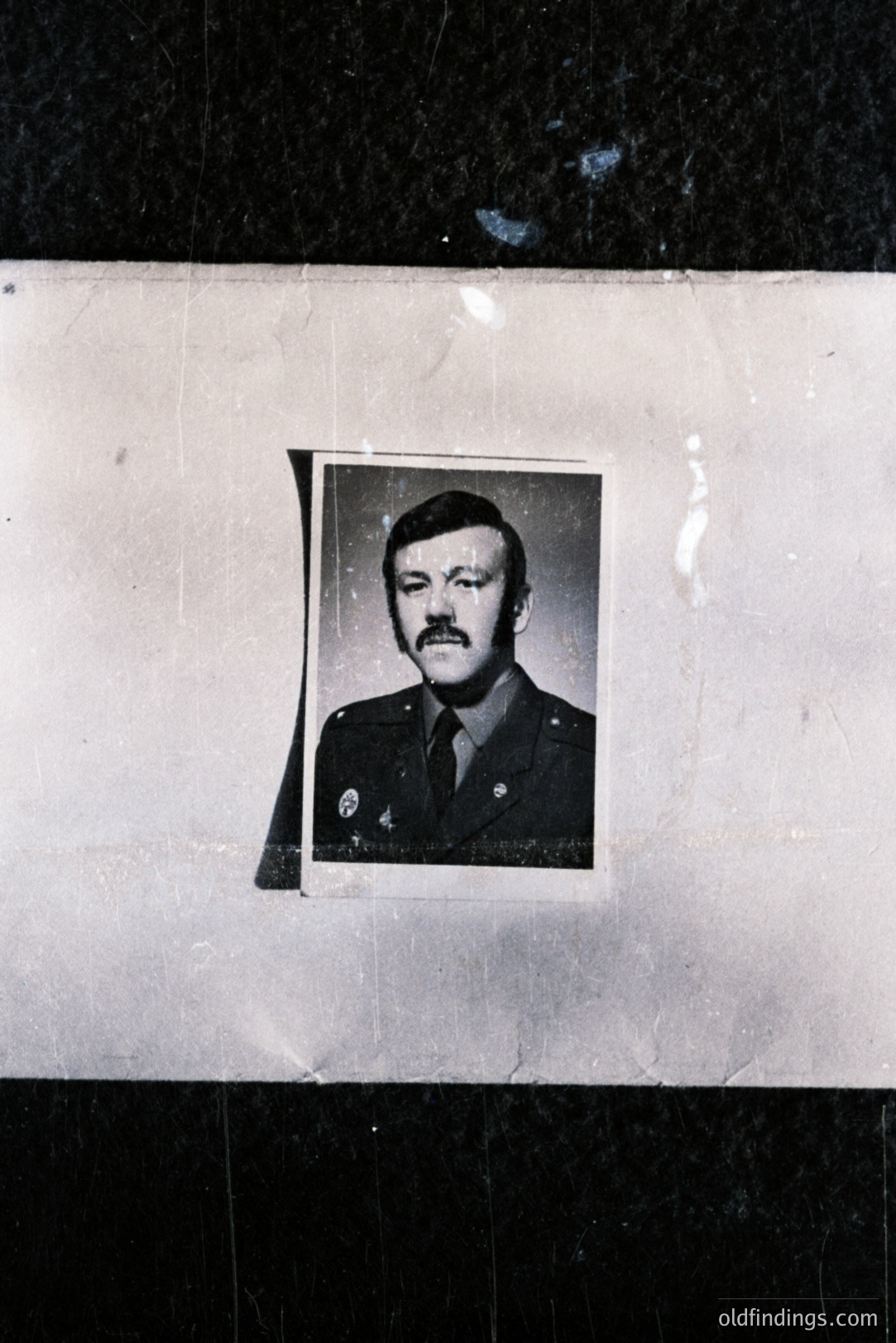 Formal portrait of a man in a military uniform, likely 1970s era, with a prominent mustache. The photograph is affixed to a textured, aged surface, showing visible wear and scratches. A striking, simple composition. Military history reference material.