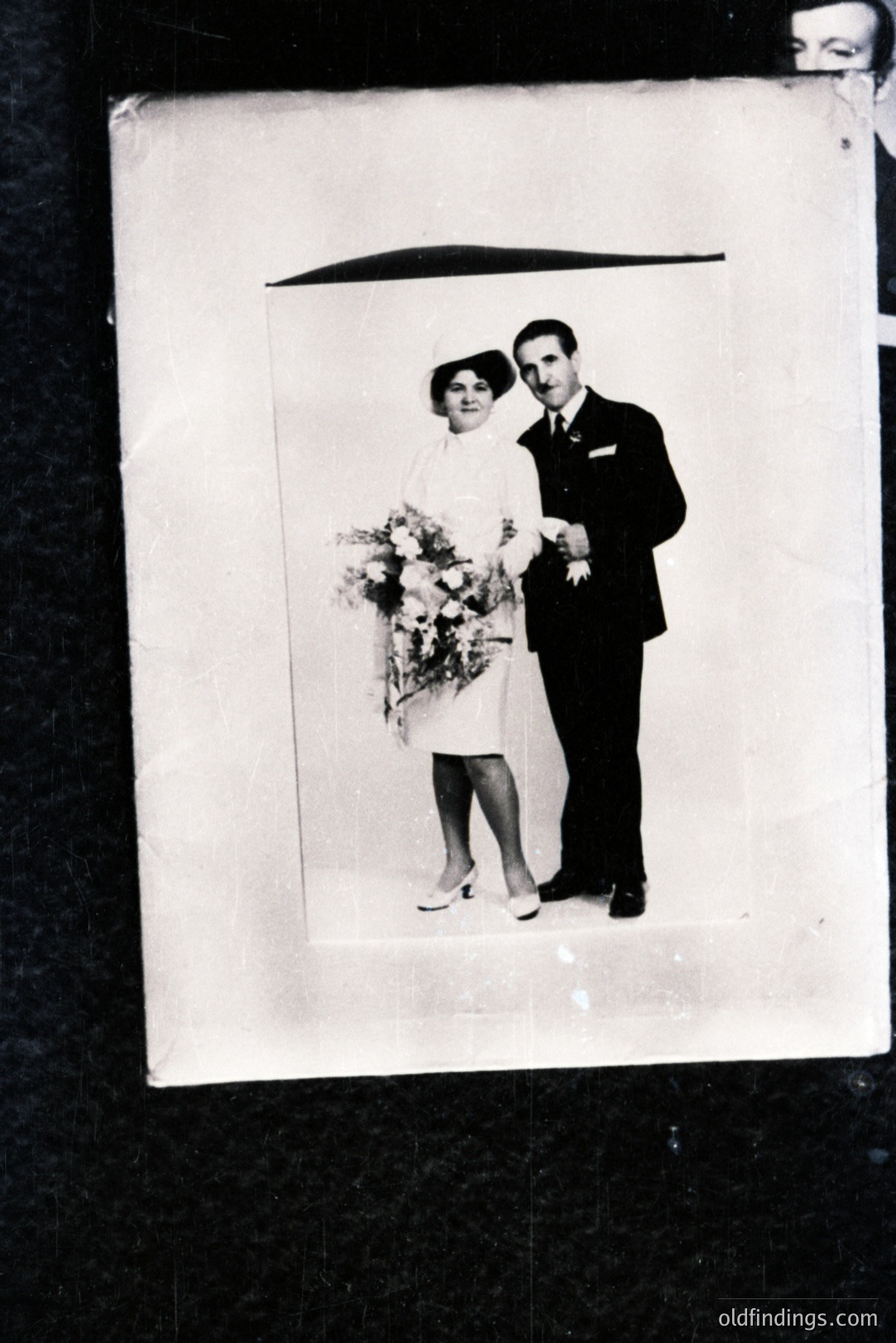 A formally posed black and white portrait of a newly married couple. The woman wears a short, fitted dress and a veiled hat, holding a bouquet of flowers. The man is in a dark suit. Likely mid-20th century, possibly 1950s-1960s. Image exhibits age-related degradation.