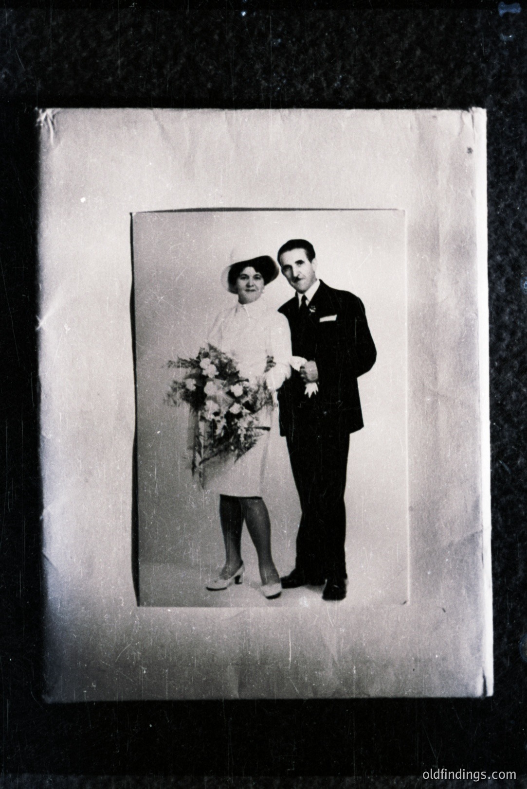 Formal studio portrait of a couple, likely a wedding. The bride wears a short-sleeved dress and carries a bouquet of flowers. Groom in a dark suit and tie. Simple backdrop, aged and marked with surface damage indicative of age. Likely 1930s-1950s. Potential for historical research or vintage design inspiration.