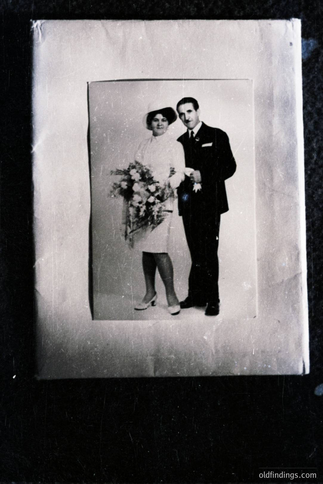 Black and white portrait of a formally dressed couple, likely a wedding photo. The woman wears a short dress and hat, carrying a bouquet of flowers. Man in a suit and tie. Likely 1950s or 1960s. Photographic print exhibits signs of age, including dust and scratches.